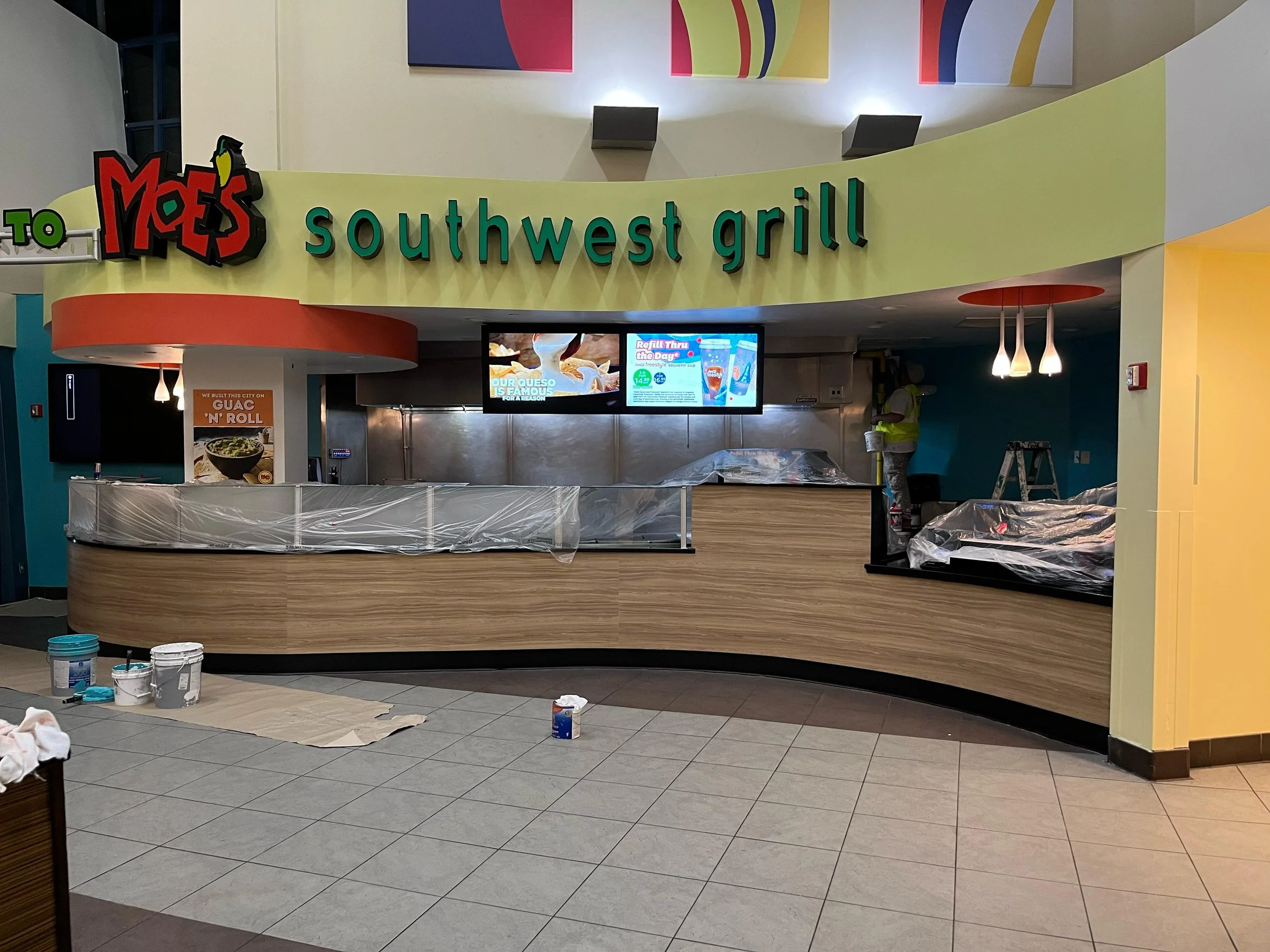 Interior of a Moe's Southwest Grill restaurant under construction, with plastic-covered counters, construction tools and paint buckets on the tiled floor, and a worker in safety vest working in the background.