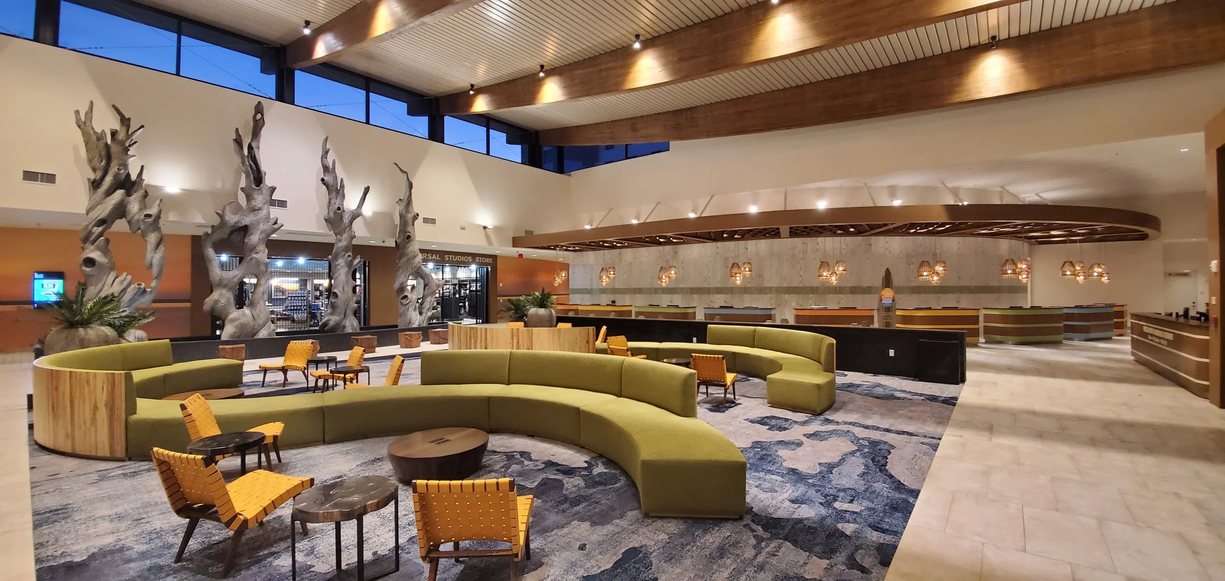 Spacious hotel lobby with large green curved sofa, wooden chairs with yellow upholstery, decorative wood sculptures resembling driftwood, potted plants, and warm lighting fixtures.