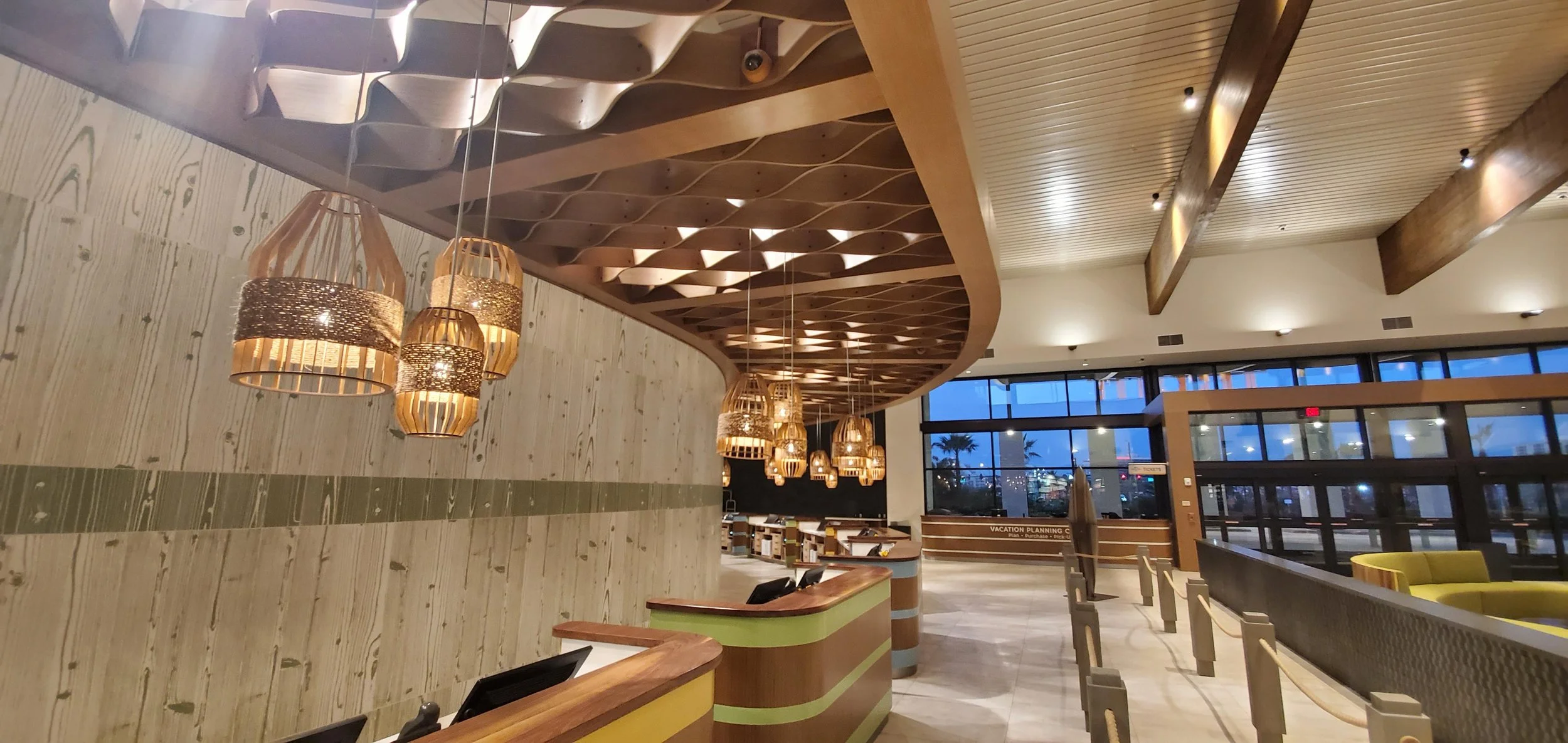 Modern hotel lobby with wooden ceiling panels, hanging wicker pendant lights, a check-in desk, and seating area with yellow chairs and large windows showing a sunset outside.