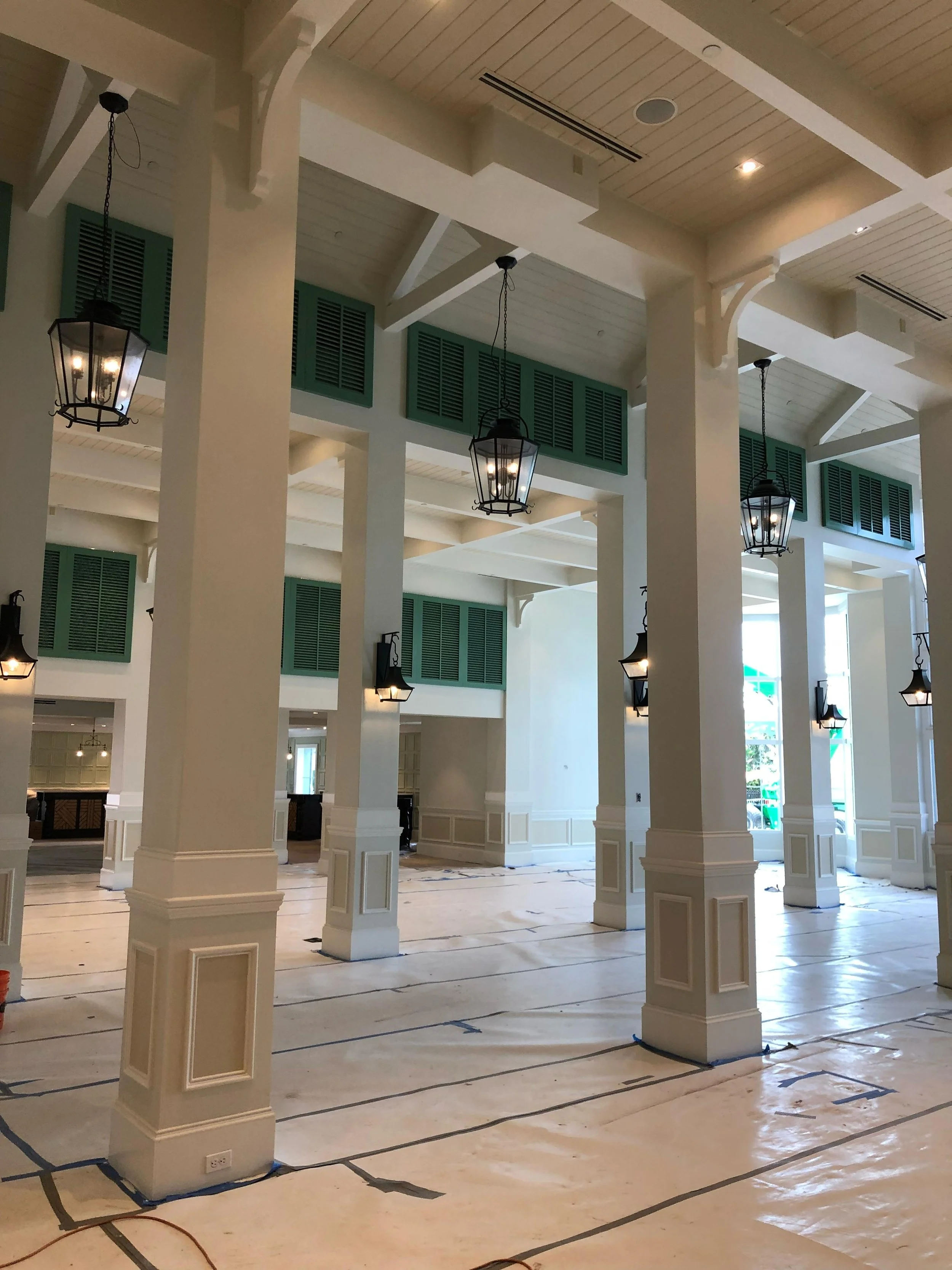 Indoor space under construction with tall white columns, green shutters, black lantern-style hanging lights, and protective floor covering.