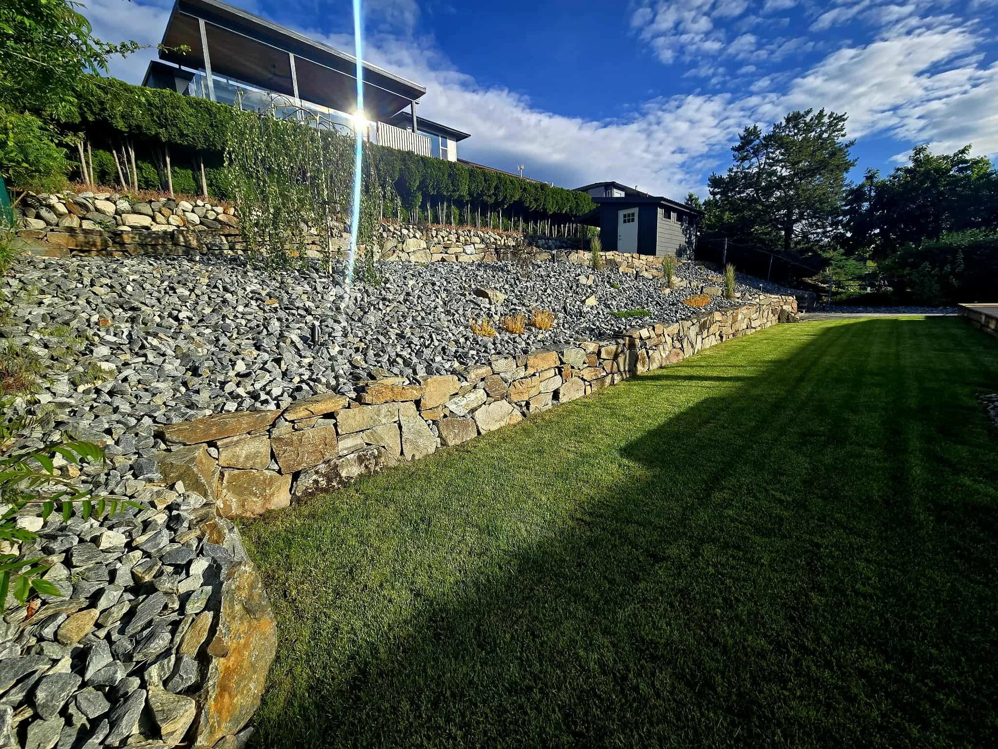 A landscaped backyard with a stone retaining wall, a small shed, a grassy lawn, and trees under a partly cloudy sky, with sunlight reflecting off the house.