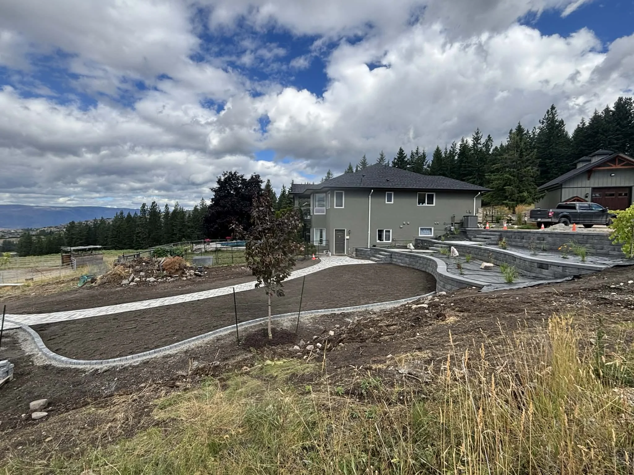 A house with a gray exterior, multiple windows, and a dark roof, situated on a hillside with a landscaped yard featuring a curved walkway, new plants, and traffic cones, with a backdrop of trees and a cloudy sky.