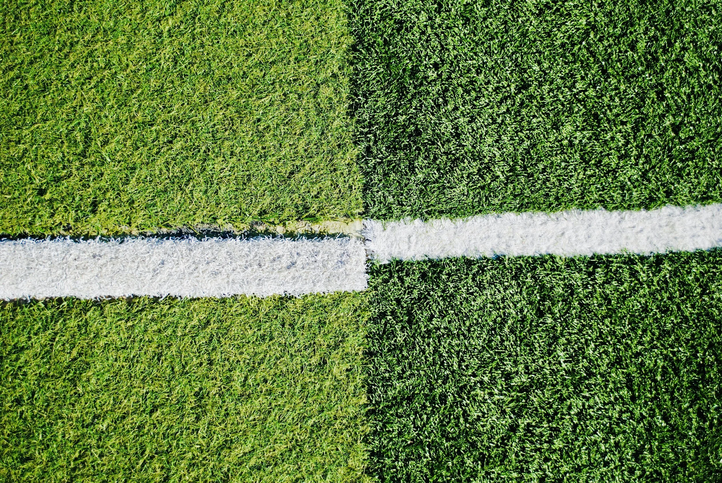A close-up aerial view of a sports field with green artificial grass, showing white boundary lines and division lines.