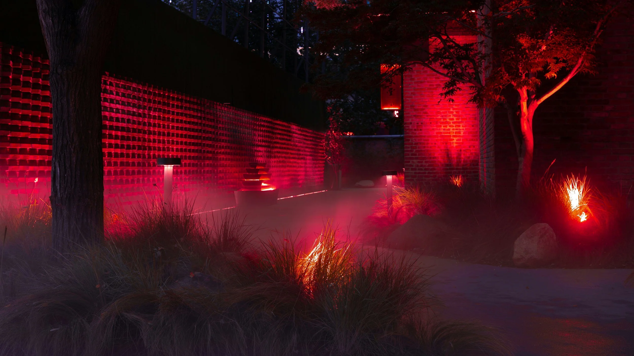 Nighttime scene of a landscaped outdoor area illuminated with red lights. There is a brown tree on the right, a brick wall on the left, and a pathway with small lights and plants along the edges.