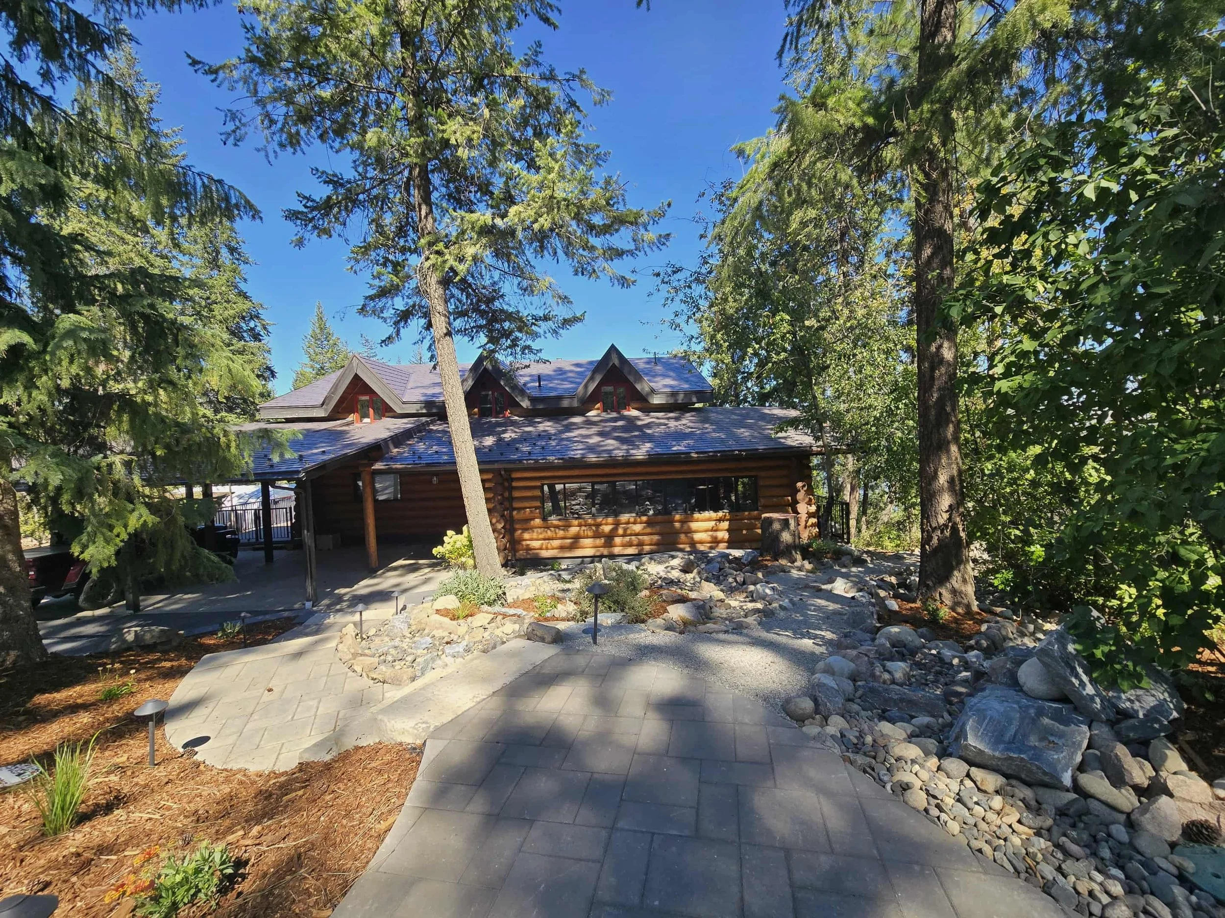 A wooden house surrounded by tall trees with a blue sky above. The house has a black roof with multiple gables, and a porch area with stairs. There is a paved pathway leading to the house and landscaped areas with rocks and small plants.
