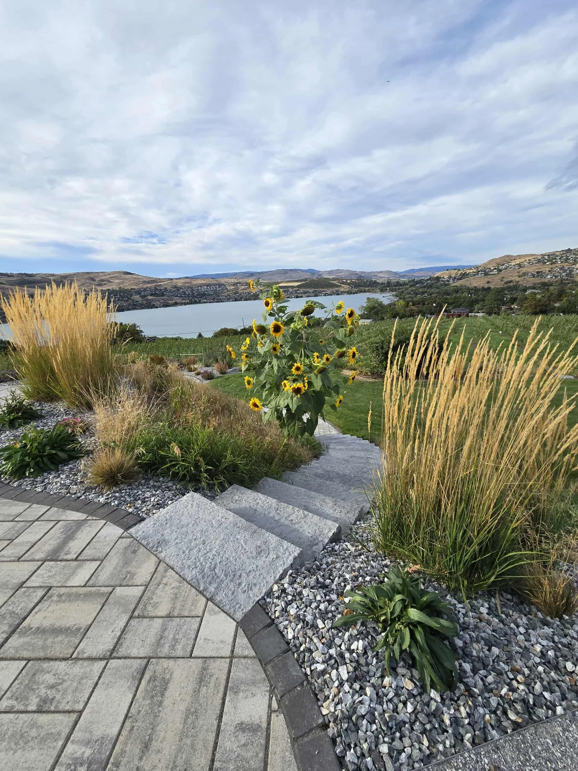 Landscape view of a large lake surrounded by hills and a partly cloudy sky, with a landscaped garden in the foreground featuring sunflowers, ornamental grasses, and paved pathways.