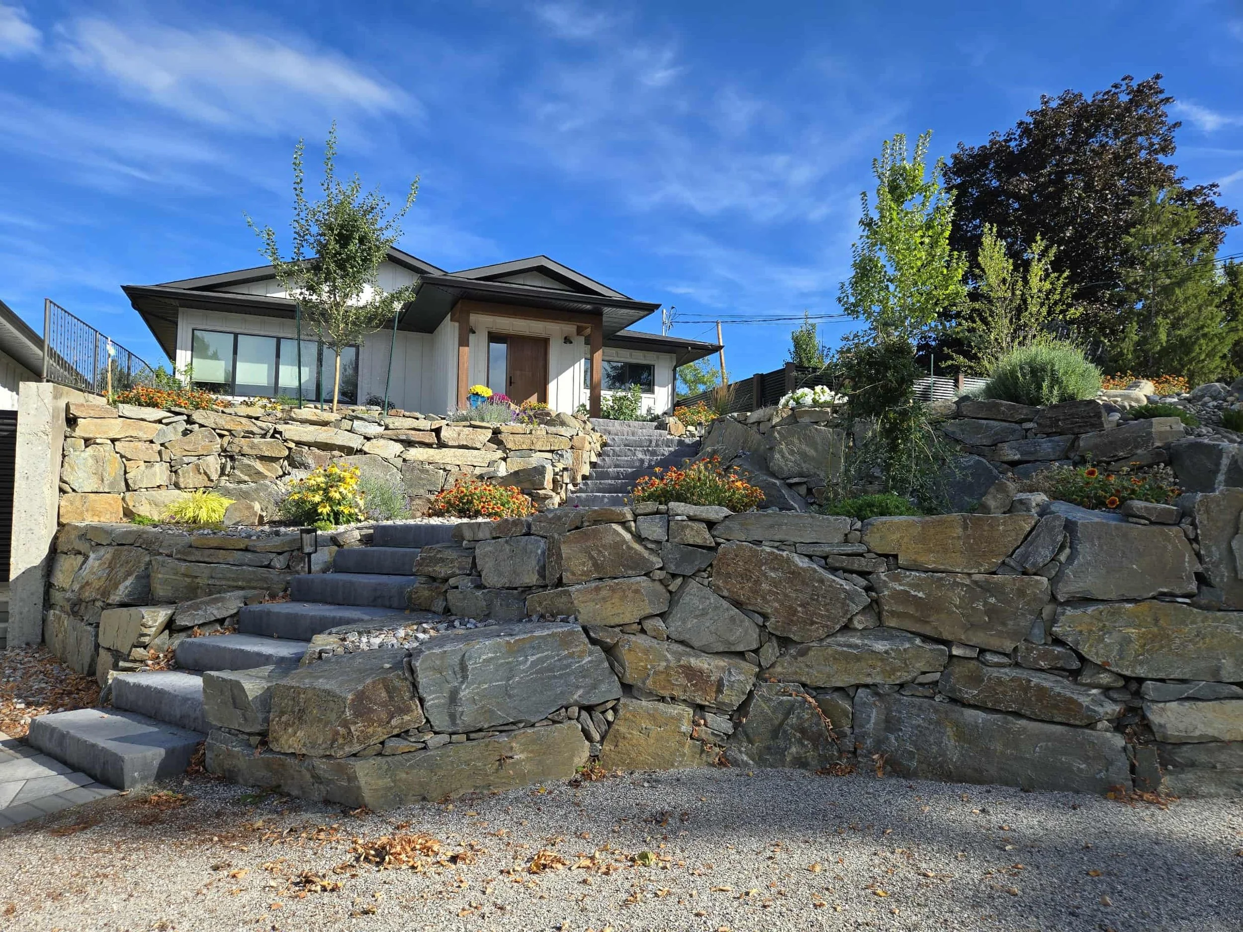 Natural stone retaining wall and staircase leading up to a modern house and surrounded by various plants and trees, located in Vernon and built by Peacock Landscaping.