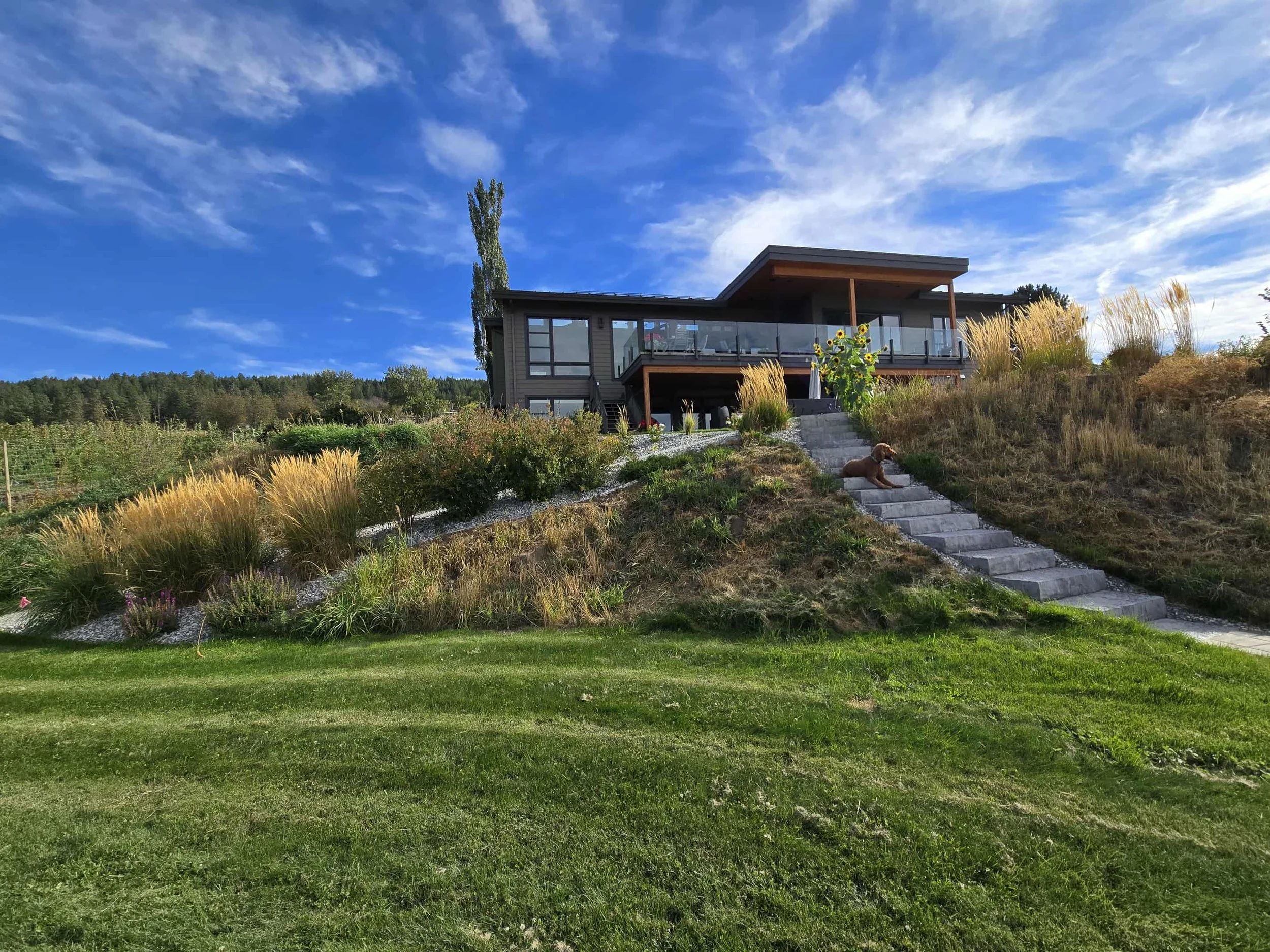 Modern house with a large balcony and glass railing, situated on a hill surrounded by grass, flowers, and trees under a partly cloudy blue sky.