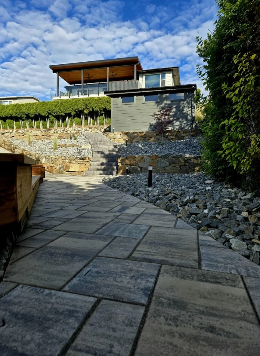A modern house with a balcony overlooking a landscaped yard with stone steps, gravel, and a paved pathway under a blue sky with clouds.