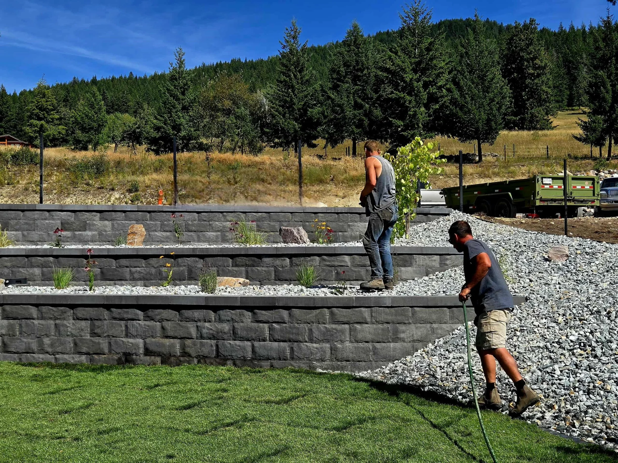 Two men working on a stone retaining wall surrounded by gravel and plants, for a residential yard in Vernon by Peacock Landscaping.
