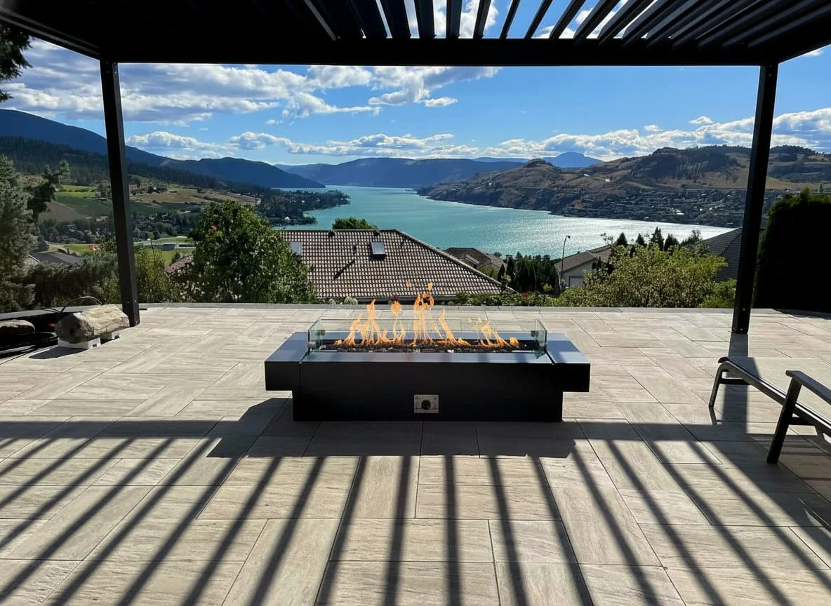 A spacious outdoor terrace with a built-in fire pit, overlooking a scenic lake and mountains in the distance on a sunny day with some clouds.
