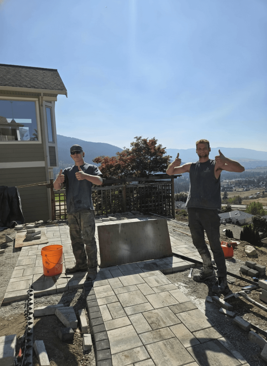 Two Peacock Landscaping crew members standing on a partially built outdoor patio in Vernon, giving thumbs up, with a view of mountains and a clear sky in the background.