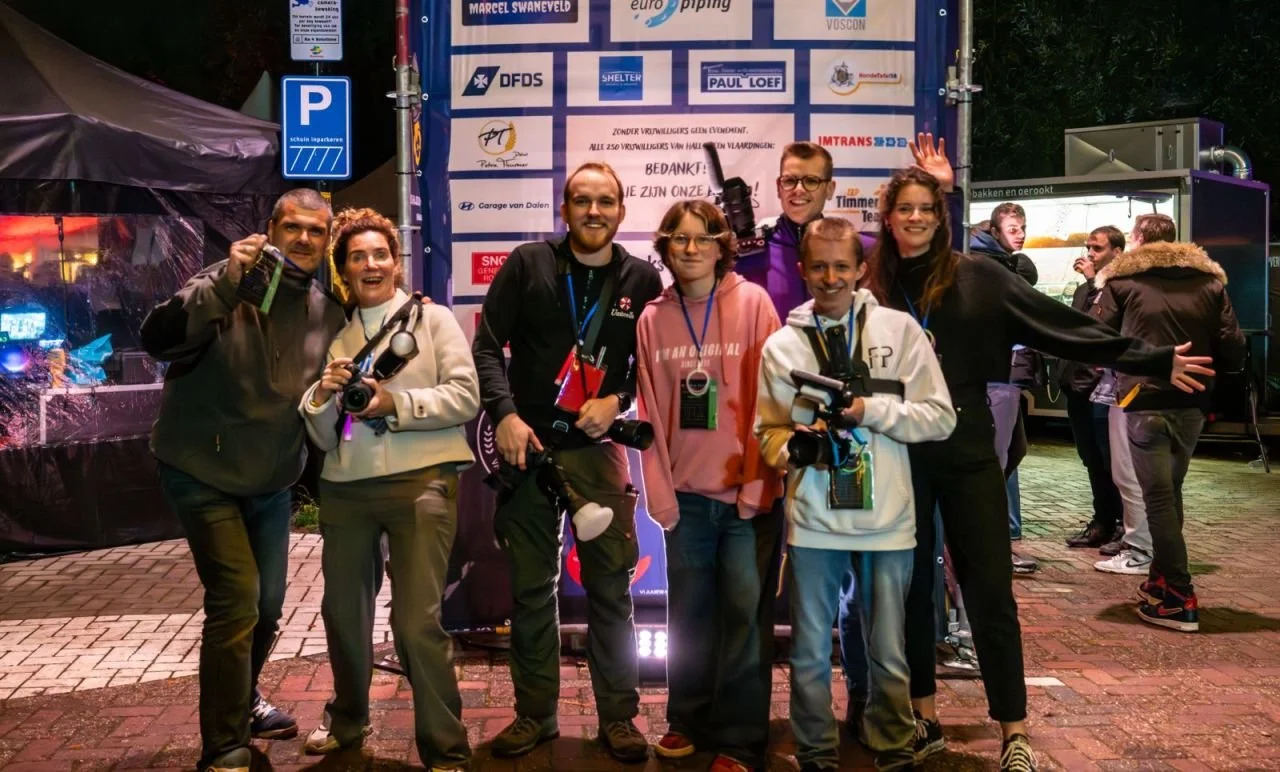 Group of seven smiling people standing in front of a sponsorship backdrop at an outdoor event, with some holding cameras and others waving.