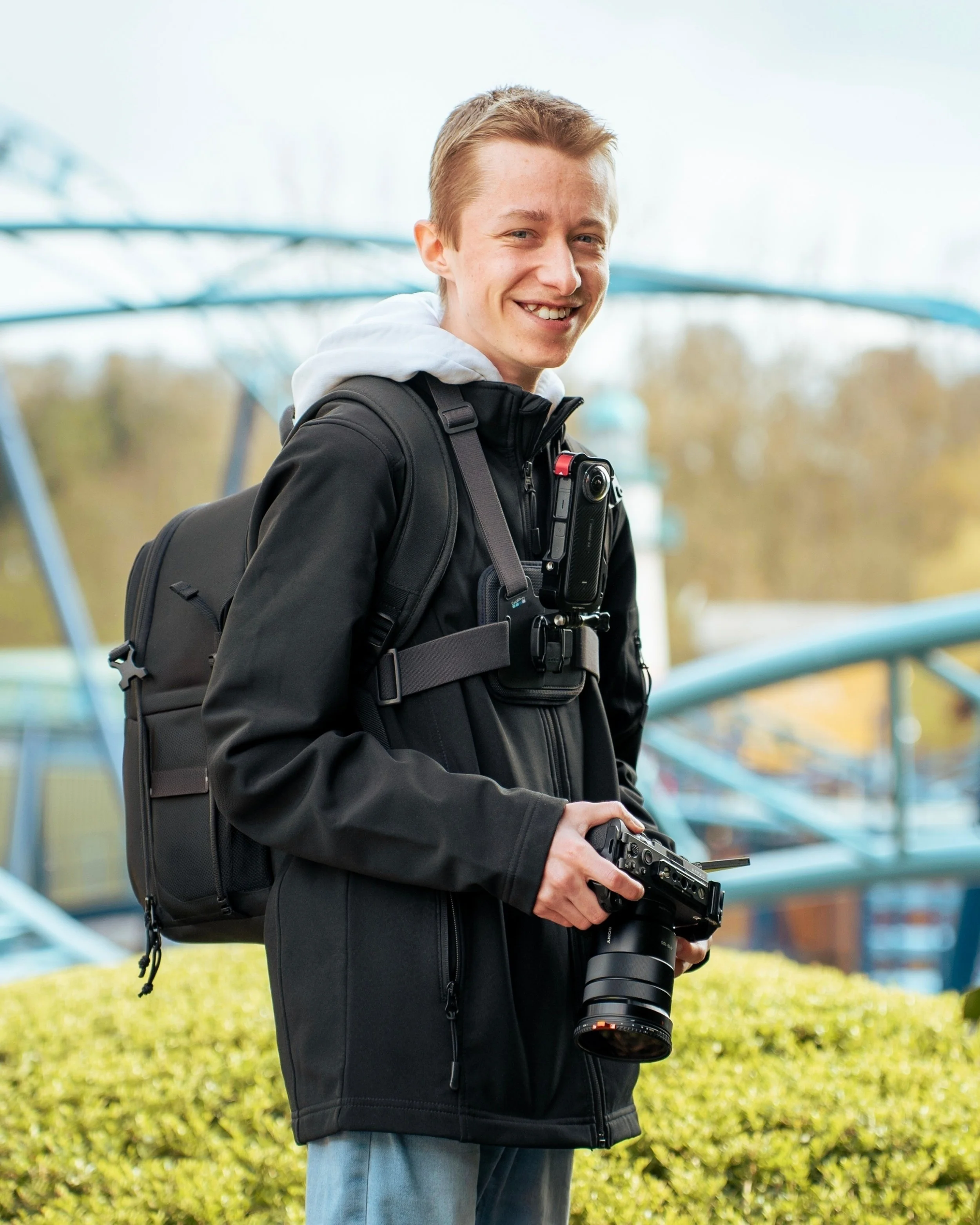 Young man standing outdoors holding a camera with a zoom lens, wearing a black jacket and a backpack, smiling.