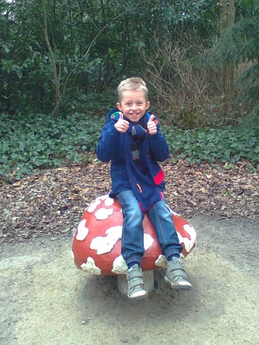 A young boy sitting on a mushroom-shaped sculpture in a wooded area, smiling and giving two thumbs up.