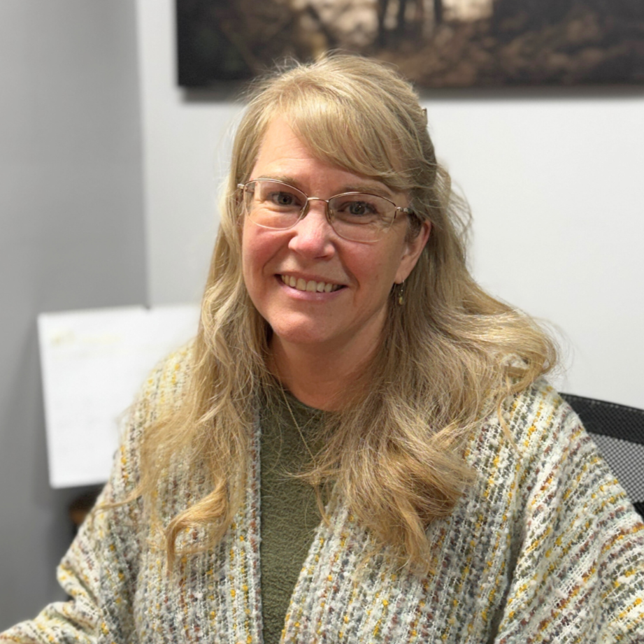 A woman with long wavy blonde hair and glasses smiling, wearing a patterned sweater, sitting in an office with a whiteboard and a framed picture in the background.