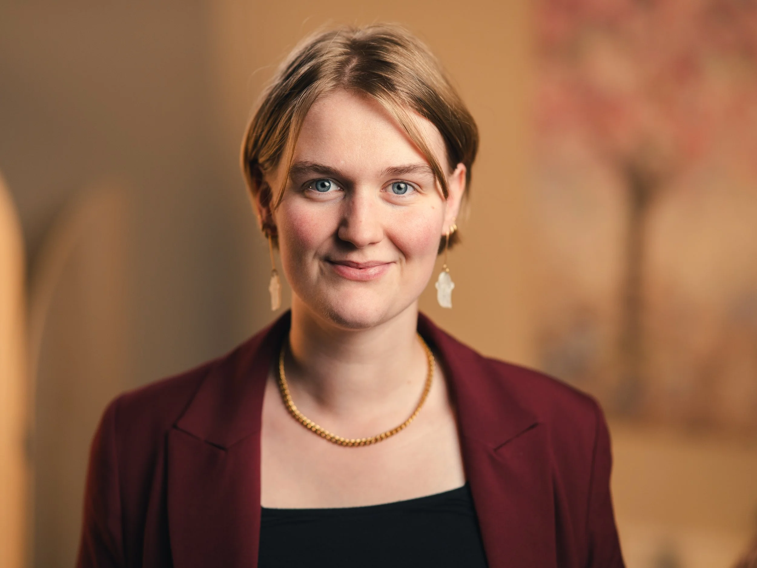 A young woman with short blonde hair and blue eyes, wearing a burgundy blazer, black top, and gold jewelry, smiling at the camera with a colorful blurred background.