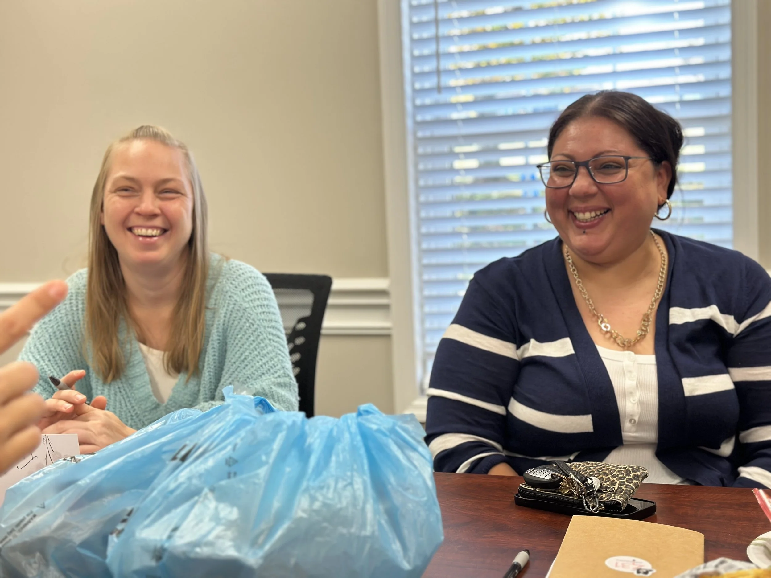Two women sitting at a table, smiling and engaging in conversation. One woman has long blonde hair, wearing a light blue sweater, and the other has short dark hair, wearing glasses, a navy blue shirt with white stripes, and jewelry. On the table are a blue plastic bag, keys, a notebook, and a pen, with a window with blinds in the background.