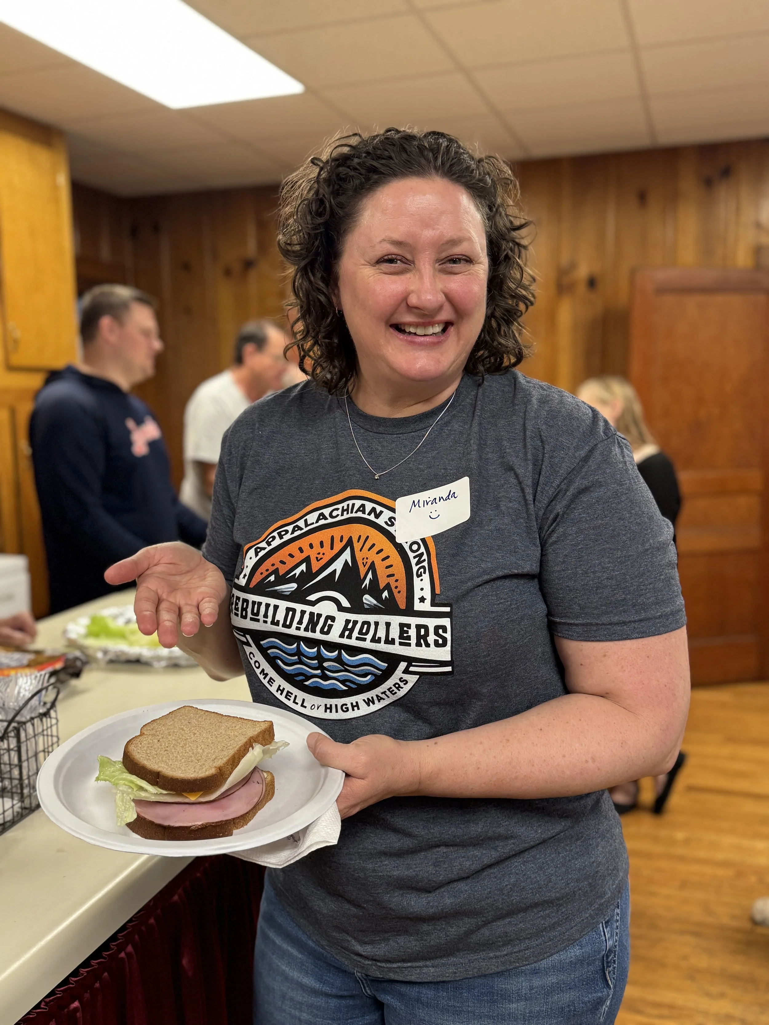 A woman smiling and holding a plate with a sandwich, wearing a gray t-shirt with a mountain and water design, in a wooden-paneled room with others in the background.