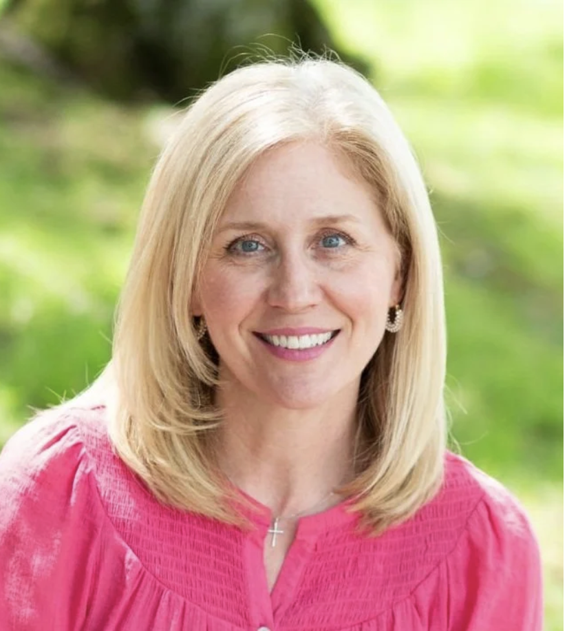 Portrait of a blonde woman with blue eyes smiling, wearing earrings, a pink top, and a cross necklace, outdoors with a green blurred background.