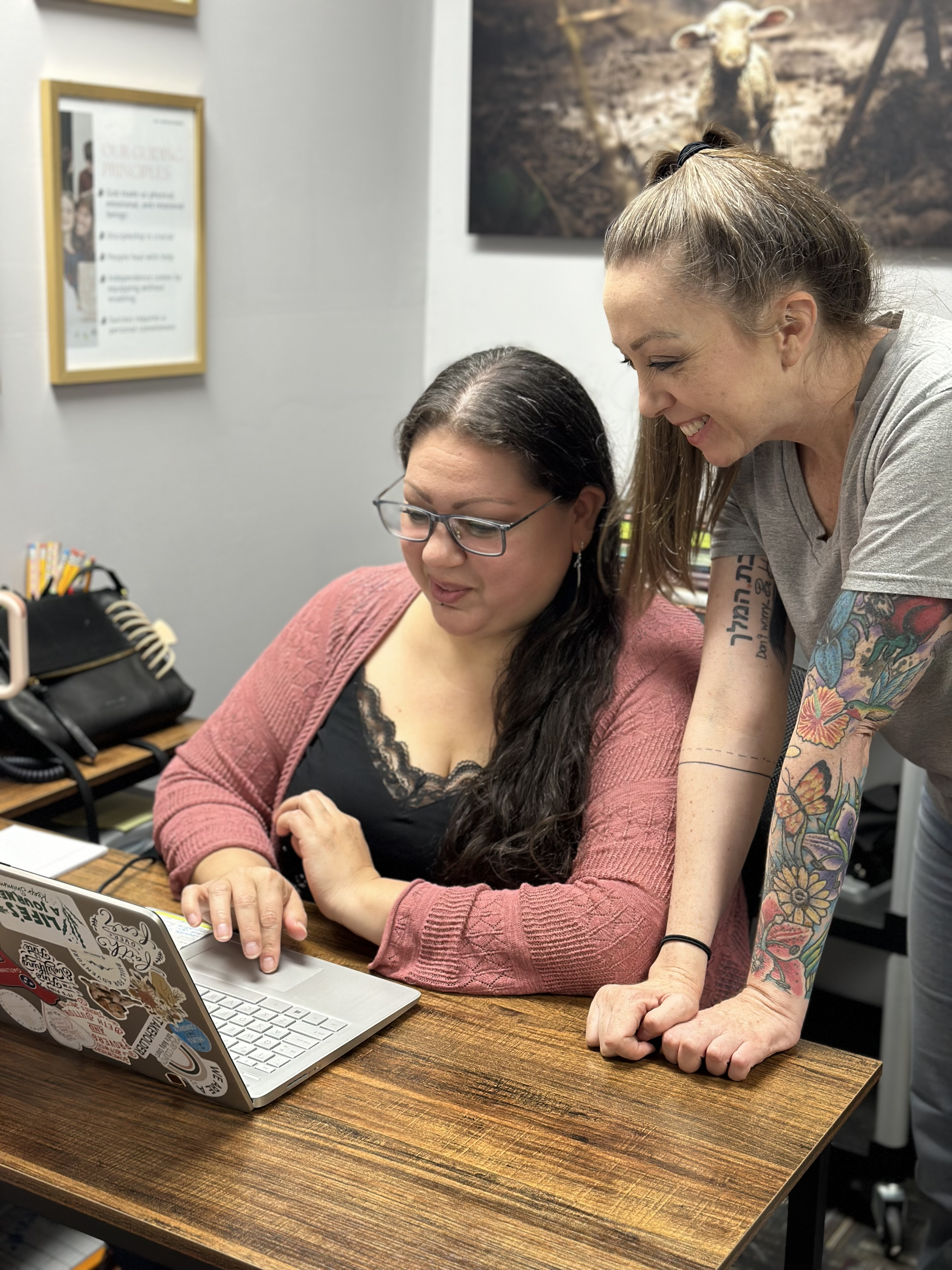 Two women sitting across from each other at a desk in an office, having a conversation. One woman is wearing a maroon top, and the other woman is wearing a gray t-shirt. The desk has office supplies, a tissue box, and beverages. There are framed pictures and a large photograph on the wall behind them.