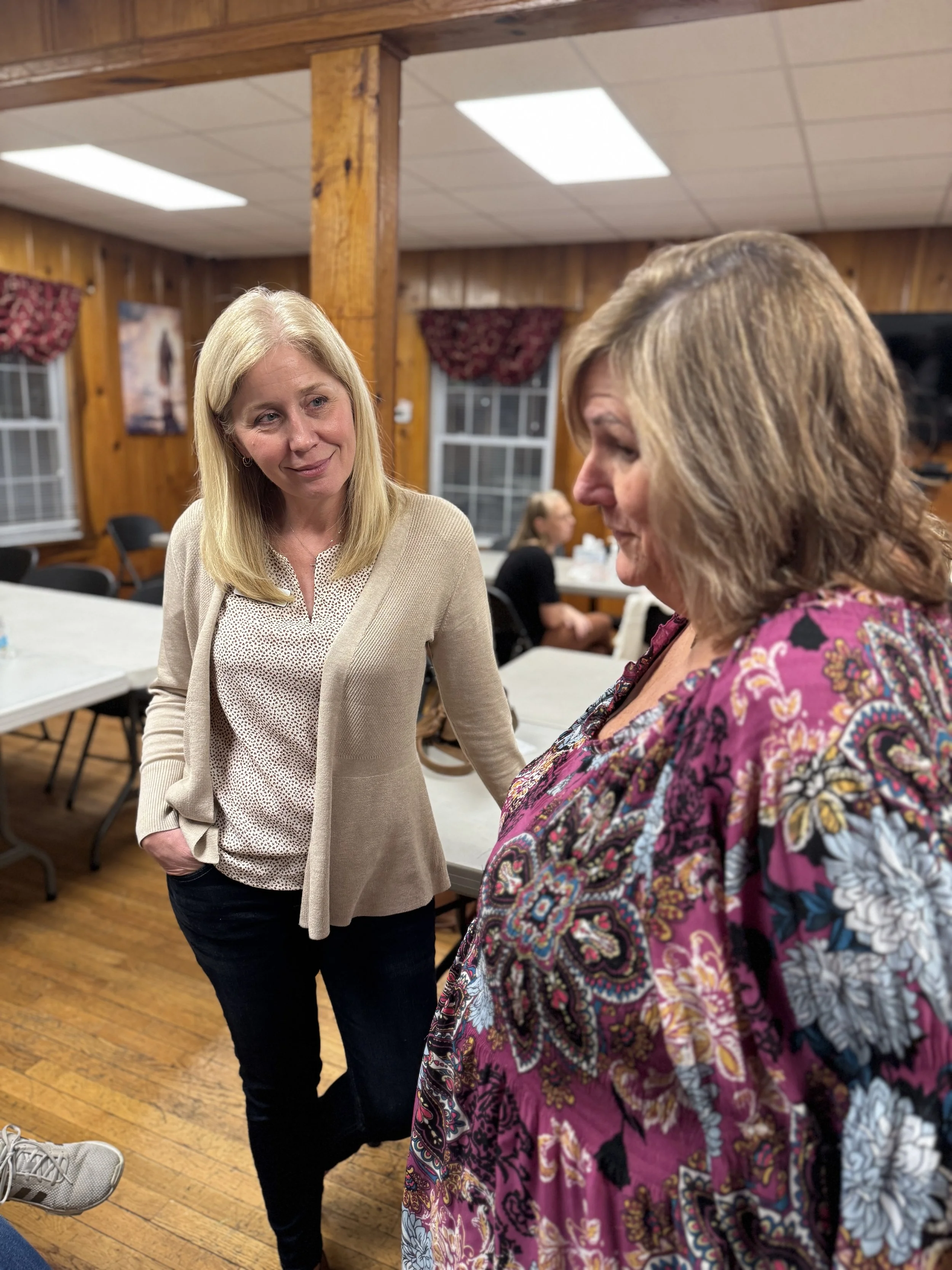 Two women engaged in conversation in a warmly lit room with wood-paneled walls and large windows. One woman with blonde hair is wearing a beige cardigan and patterned blouse, while the other woman, with light brown hair, is wearing a colorful floral top. Several tables and chairs are visible in the background.
