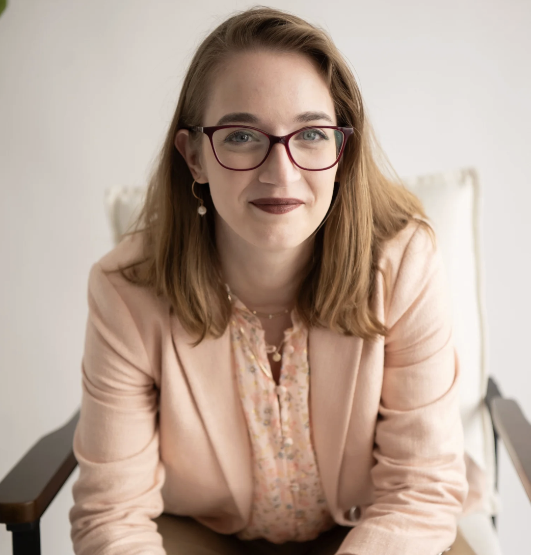 A woman with red hair, glasses, and dark lipstick sitting in a light-colored room