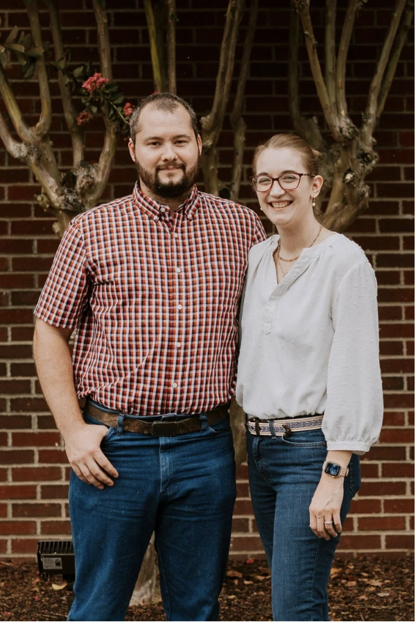 A man and woman standing outdoors in front of a brick wall and tree with pink flowers. The man is wearing a red checkered shirt and blue jeans, and the woman is wearing glasses, a white blouse, and dark jeans.