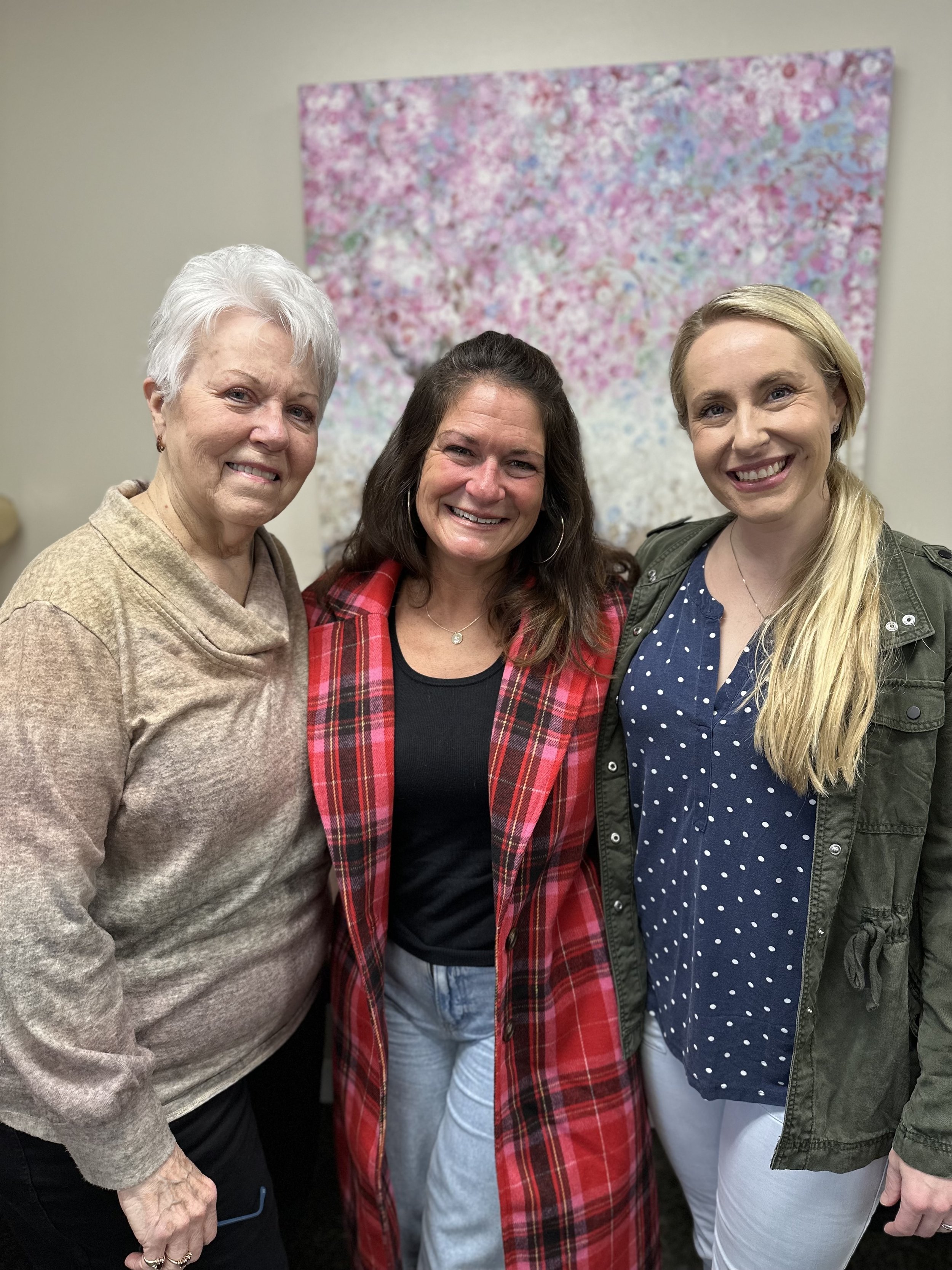 Three women smiling and standing close together in front of a colorful floral painting.