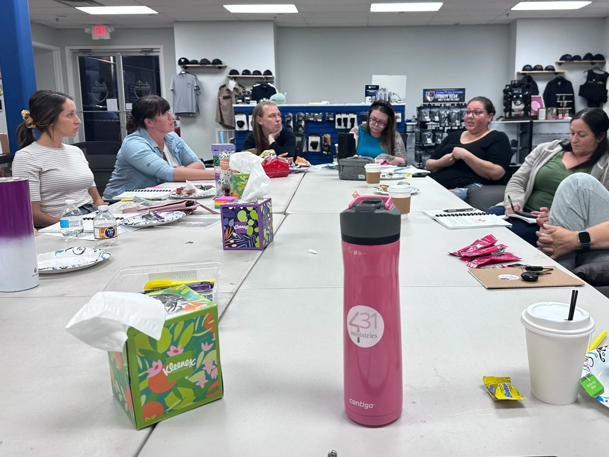 Meeting of eight women seated around a long conference table with snacks, drinks, and notebooks, in a retail store or office setting.