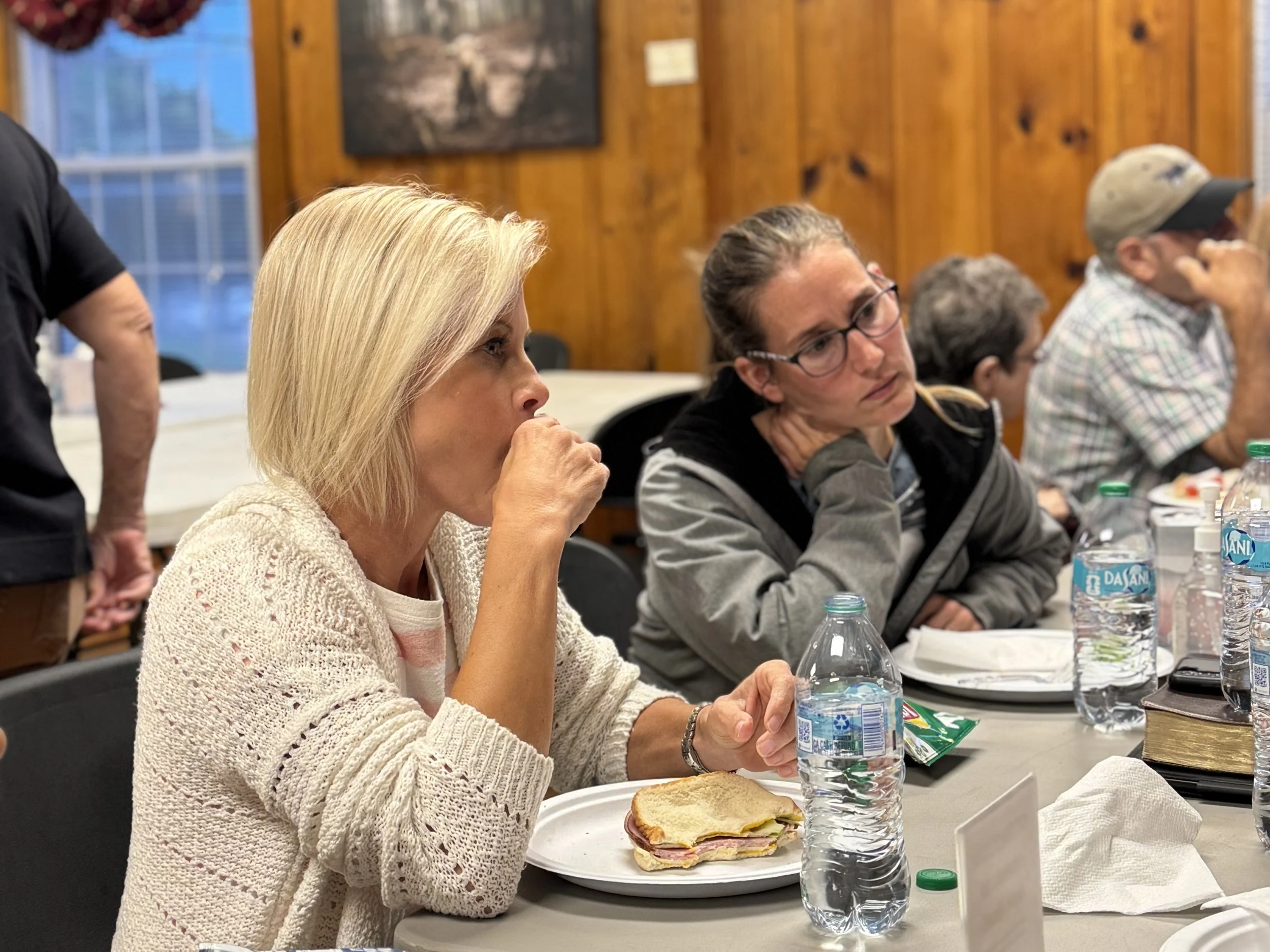 A woman with blonde hair wearing a beige knit sweater eating a sandwich at a table with bottled water and other people in the background.