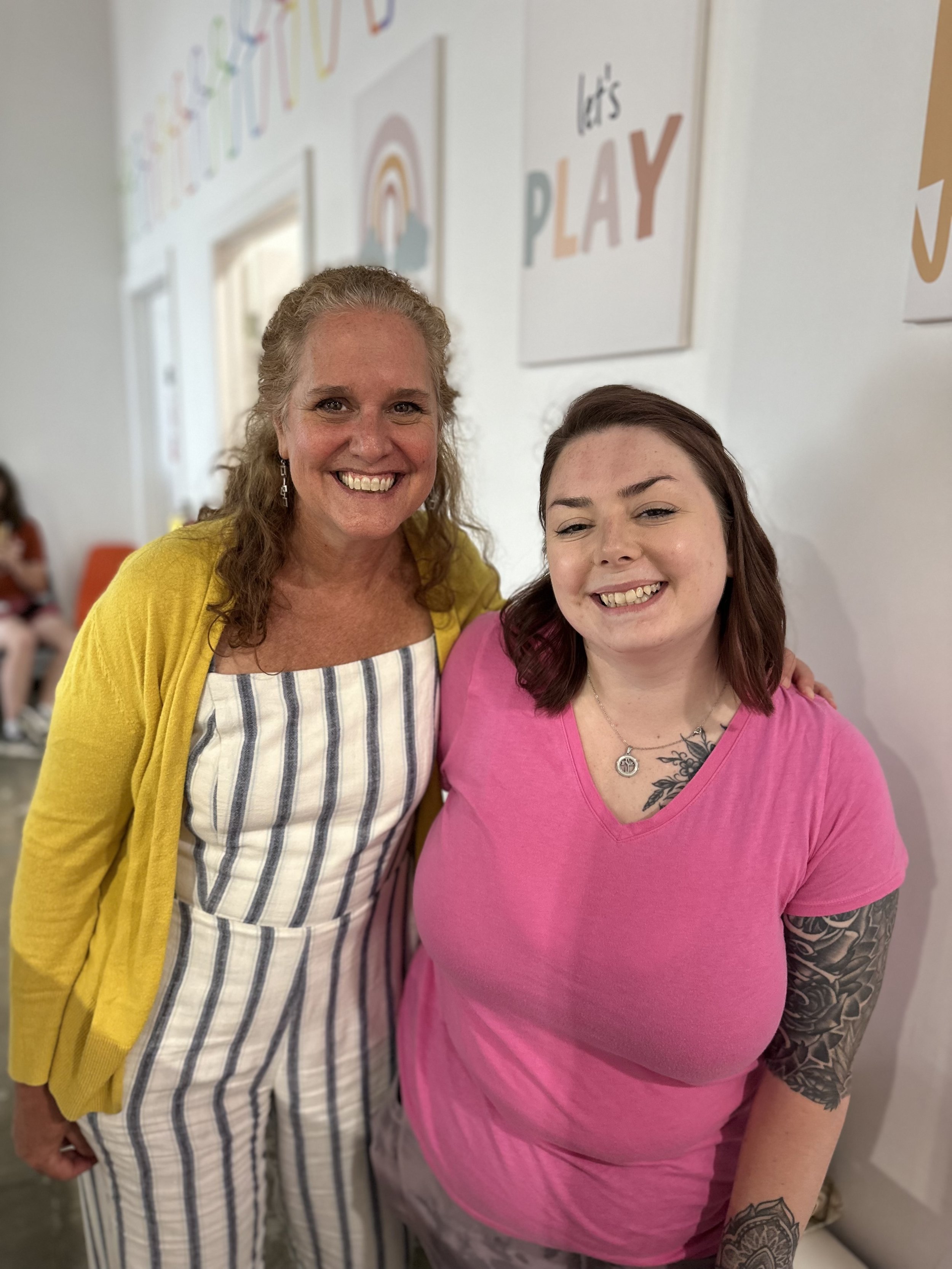 Two women smiling, standing close together indoors, with colorful wall art behind them that says "Let's Play." One woman is wearing a yellow cardigan and striped jumpsuit, and the other woman is wearing a pink t-shirt with tattoos on her arm.