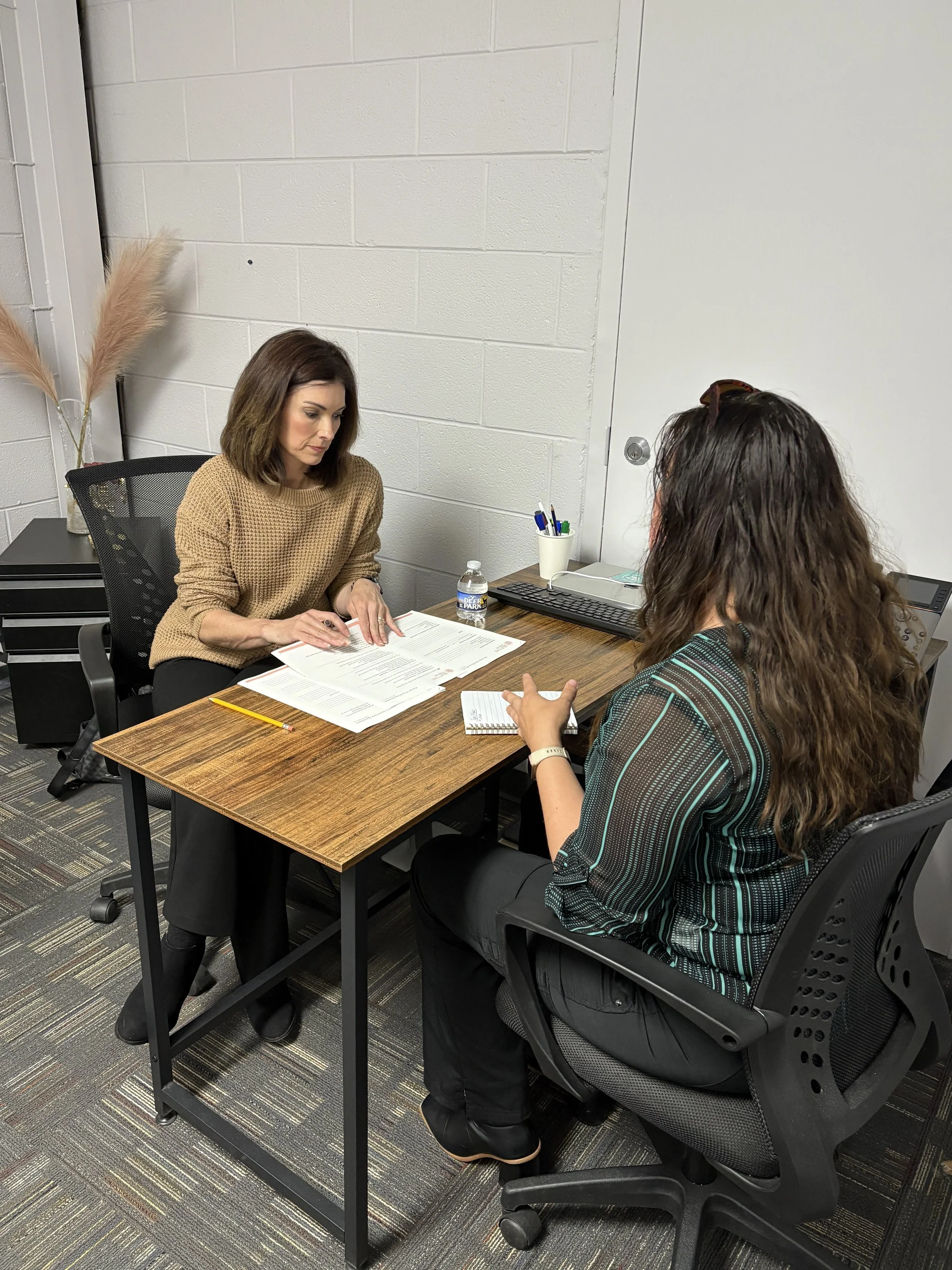 Two women having a meeting at a small office desk, with papers, pens, and a water bottle.