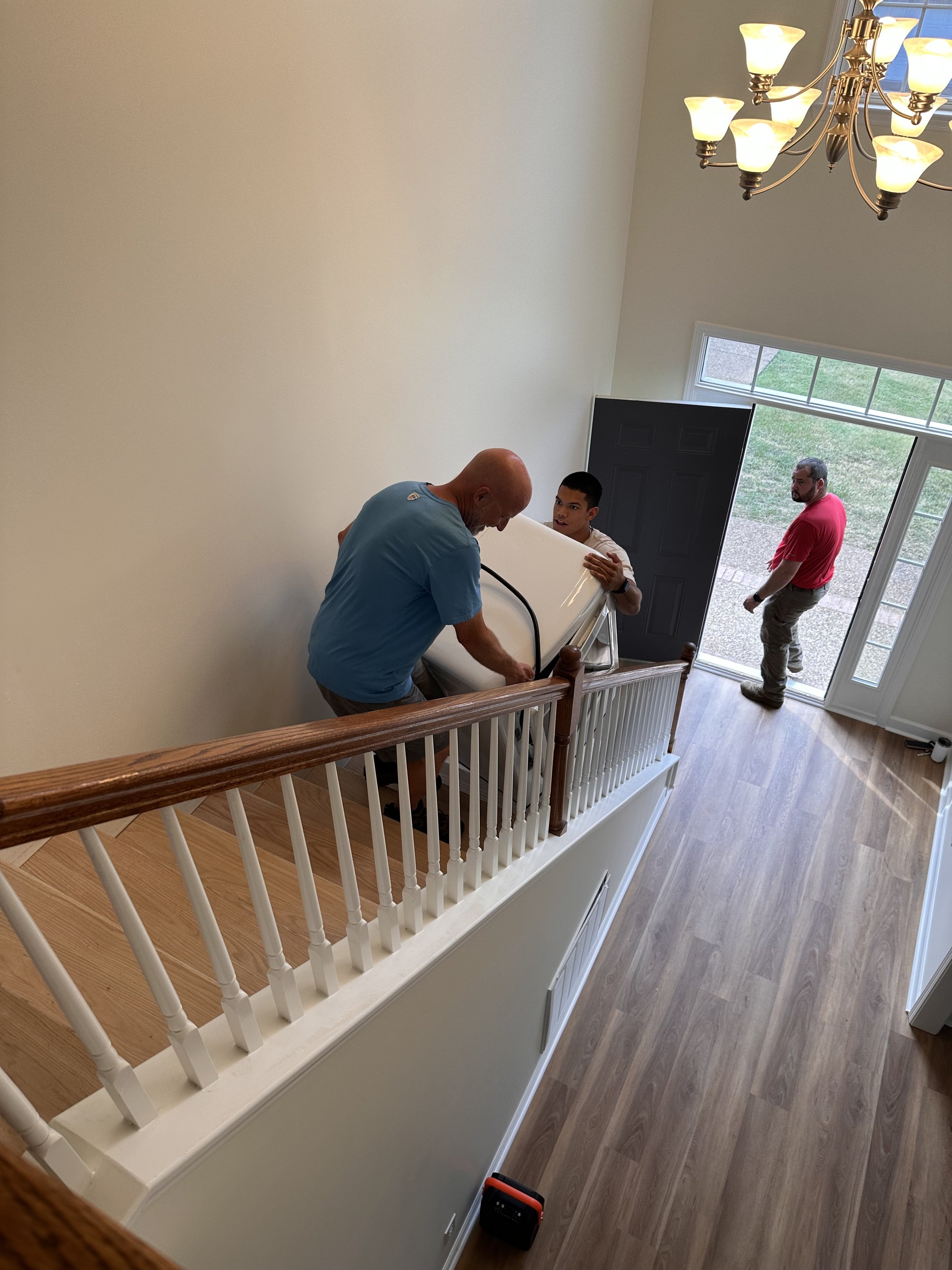 Three men are carrying a white appliance up a staircase inside a house with hardwood flooring and a large window near the front door.