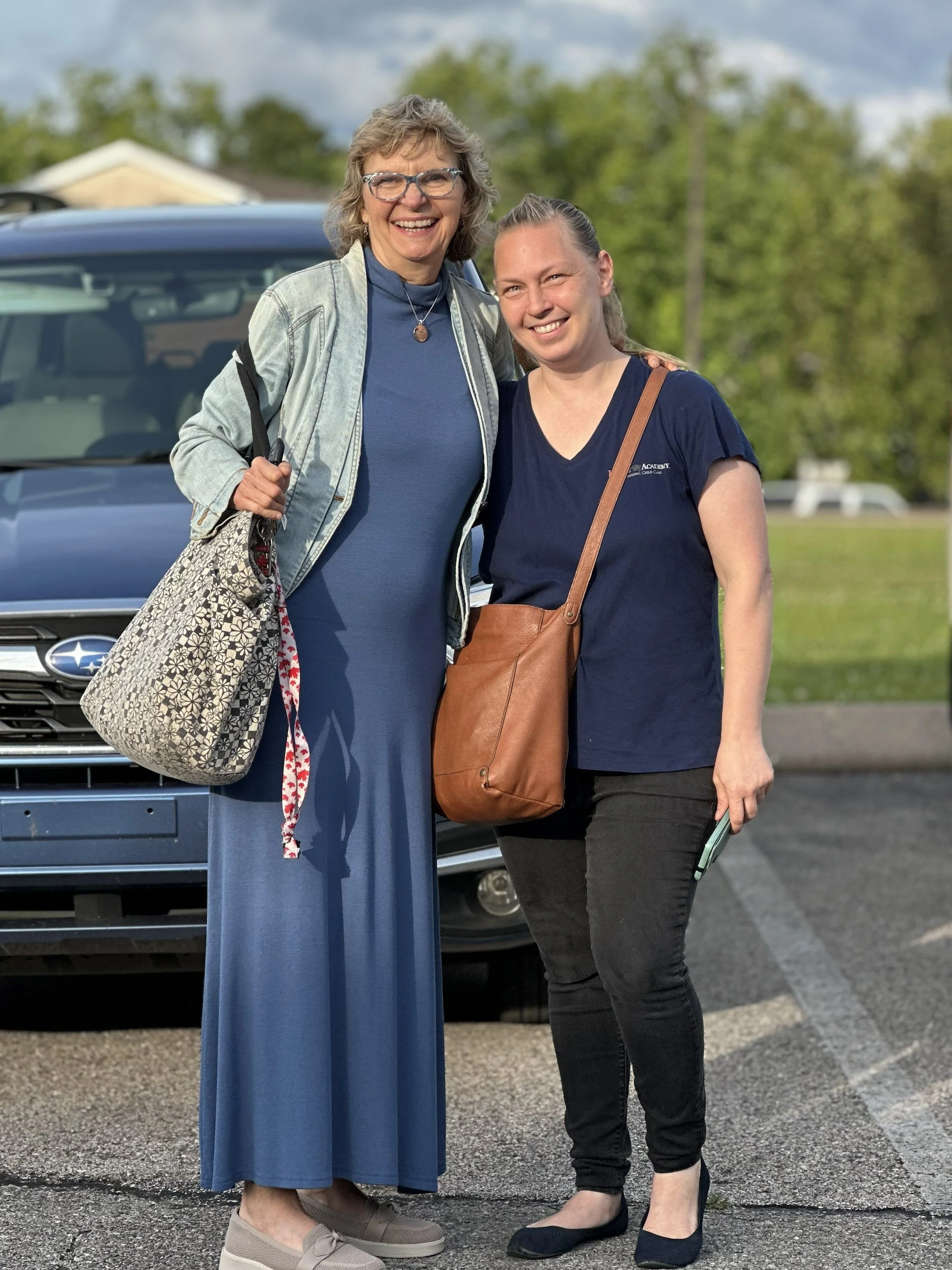 Two women standing in a parking lot, smiling, with a car behind them and trees in the background.