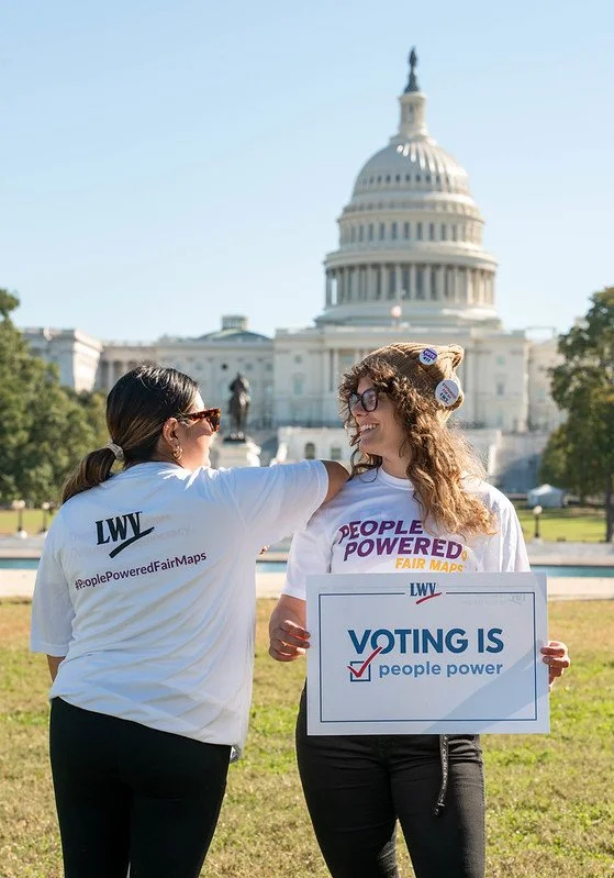 Two women protesting for voting rights in front of the U.S. Capitol. One woman wears a shirt with #PeoplePoweredFairMaps and the other holds a sign that says "Voting is people power."