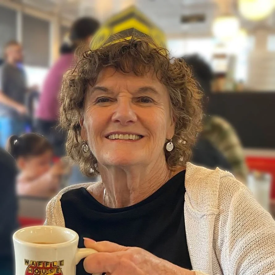 Smiling woman with curly hair enjoying a cup of coffee or tea at a restaurant, with a blurred background of other people.