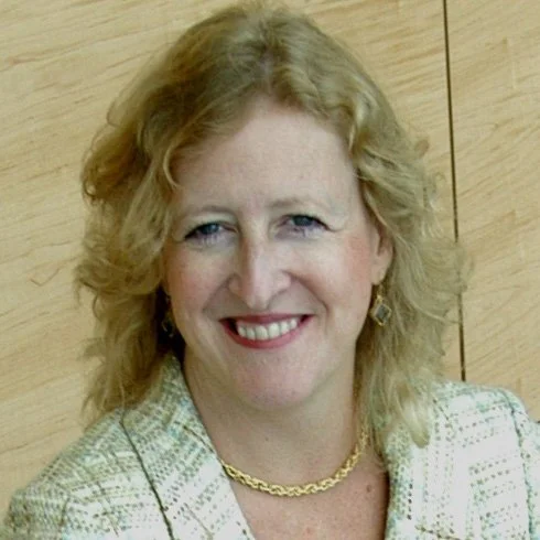 Portrait of a smiling woman with blonde curly hair, wearing a light-colored blazer, gold necklace, and earrings, standing against a wooden background.