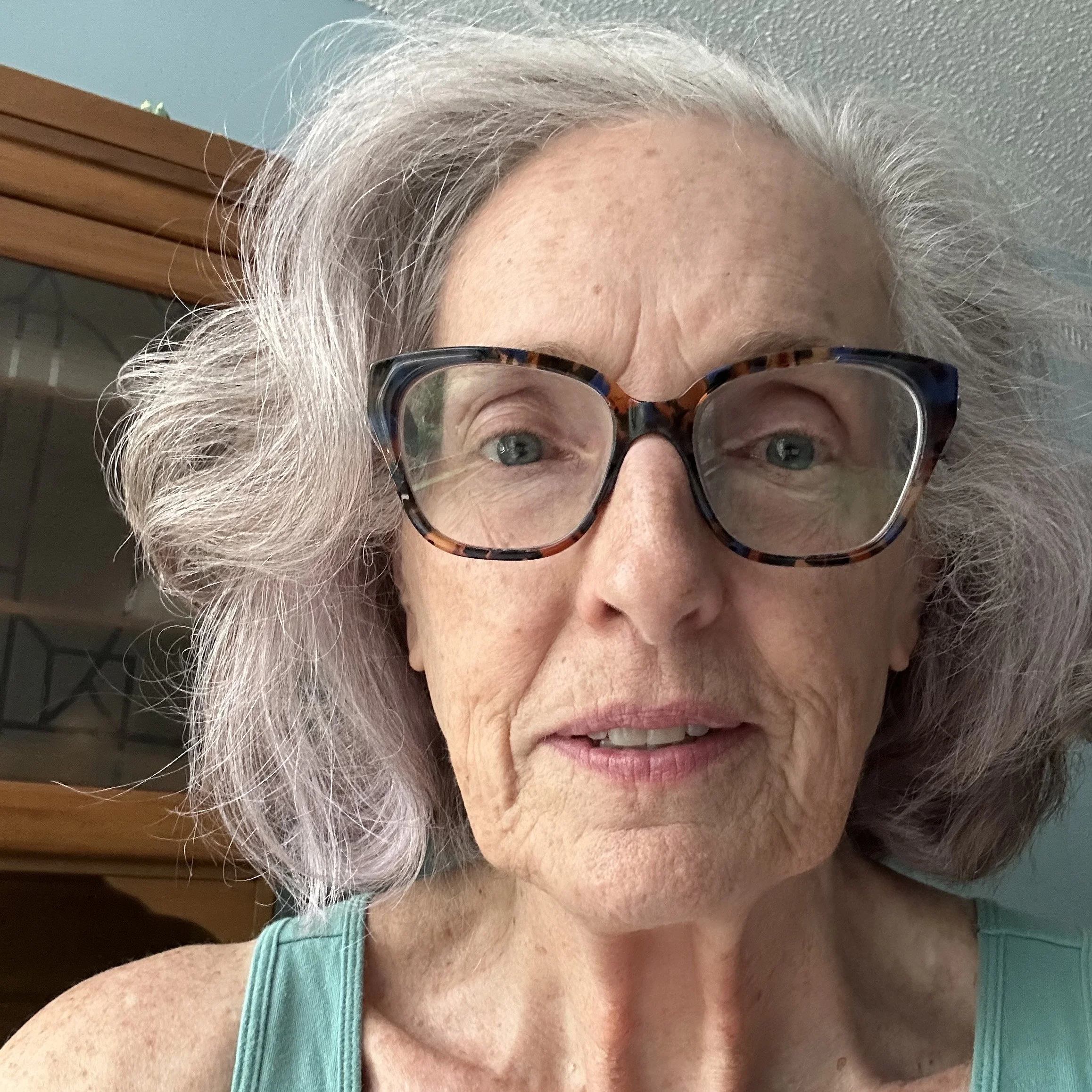 Close-up of elderly woman with gray hair, wearing large tortoiseshell glasses and a turquoise tank top, indoors with a wooden cabinet in the background.