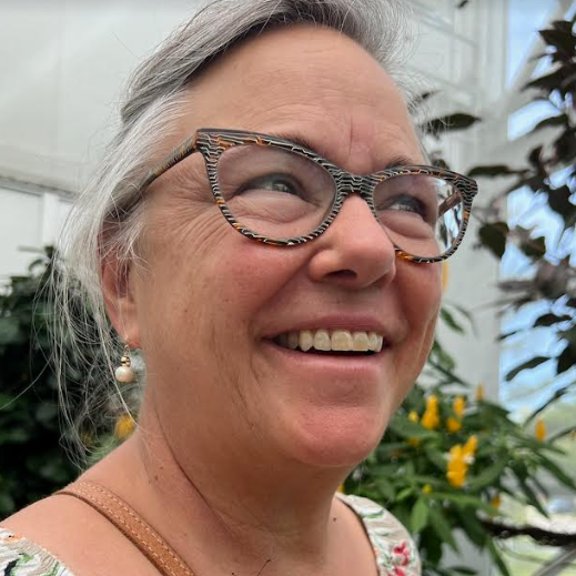 Close-up of a smiling woman with glasses in a greenhouse surrounded by plants and yellow flowers.