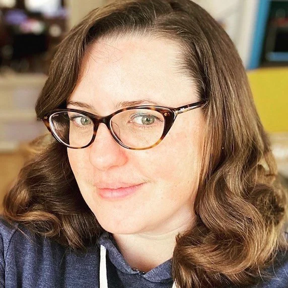 A woman with wavy brown hair wearing glasses and a navy hoodie, smiling slightly.