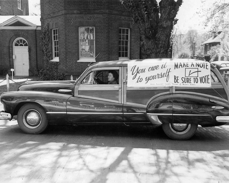 Vintage car parked on a street with a street sign in the background, and a large banner on the side of the car reading 'You owe it to yourself. Make a note. Be sure to vote.'