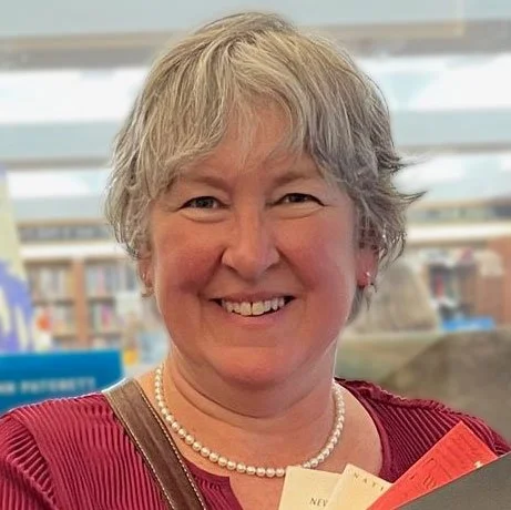 A smiling woman with short gray hair, wearing a pearl necklace and a red top, standing indoors with bookshelves in the background.