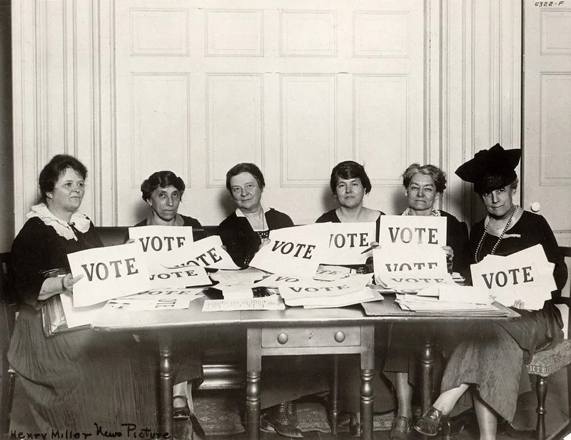 Six women sitting at a table holding signs that say "VOTE" in an old black and white photograph.