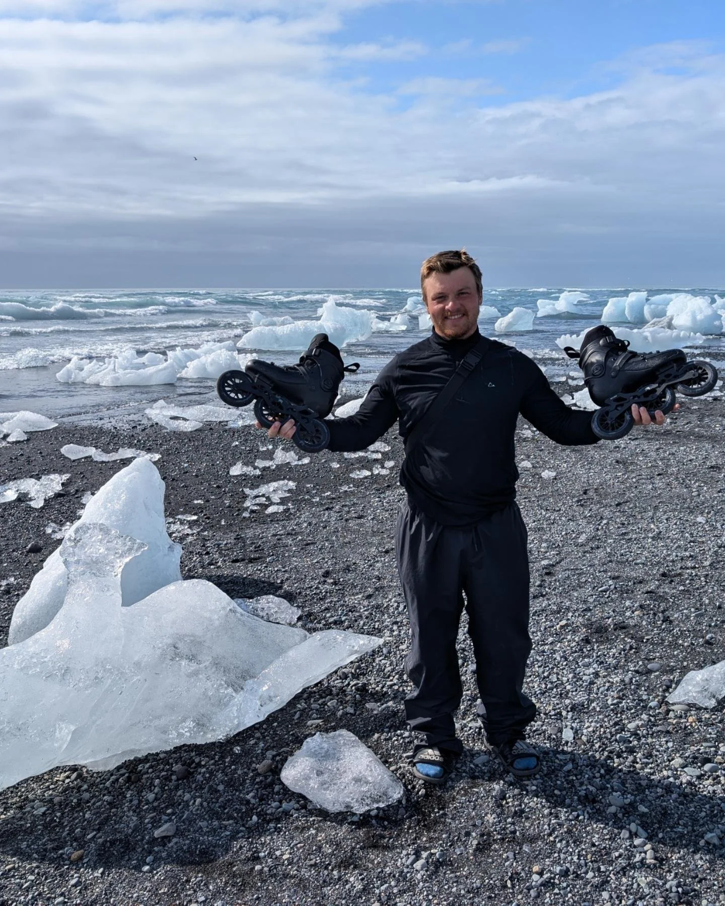 A man in black clothing holding ice skates, standing on a rocky beach with ice chunks and ocean waves in the background.