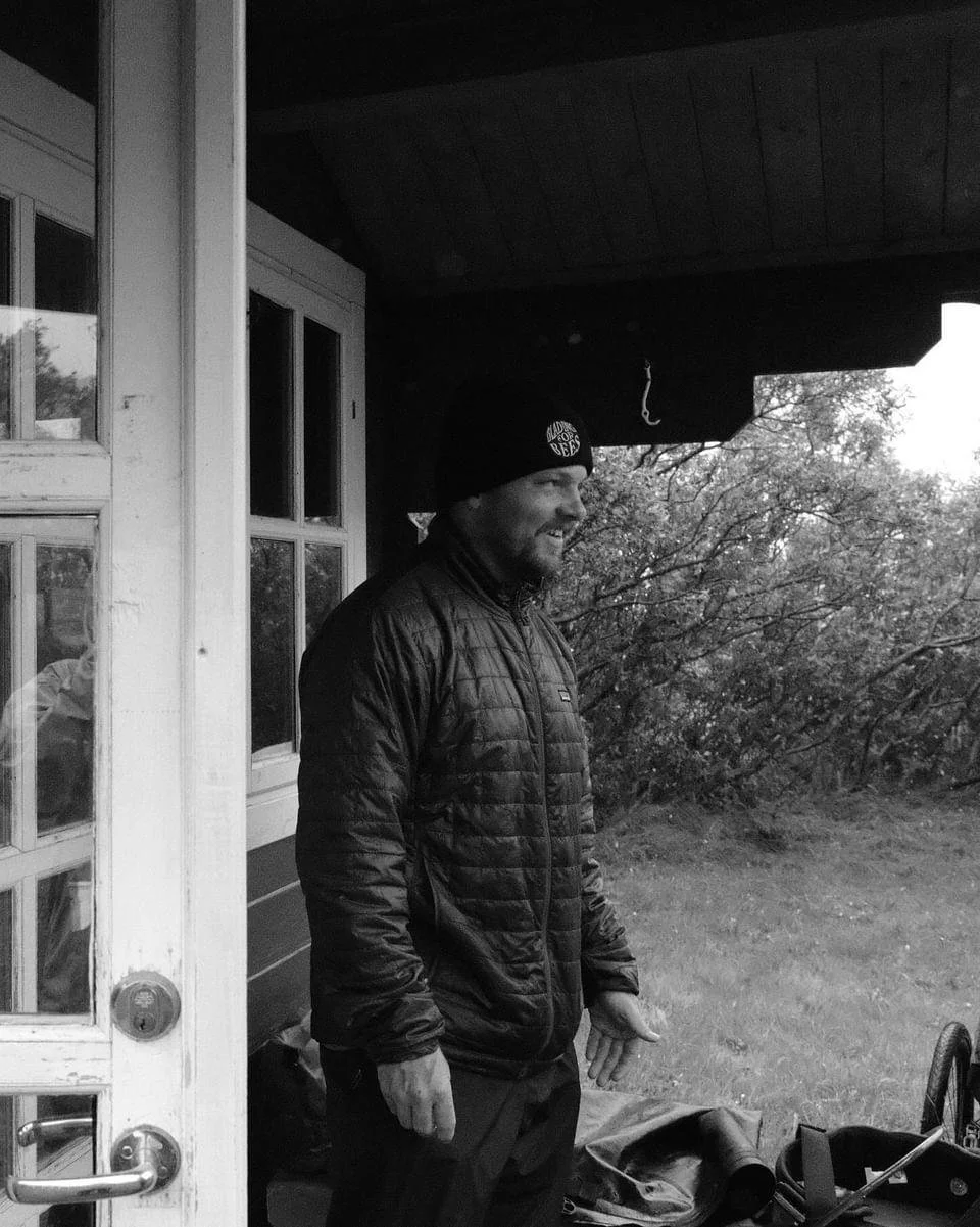 A man wearing a beanie and a quilted jacket standing outside a house, smiling, with trees in the background.