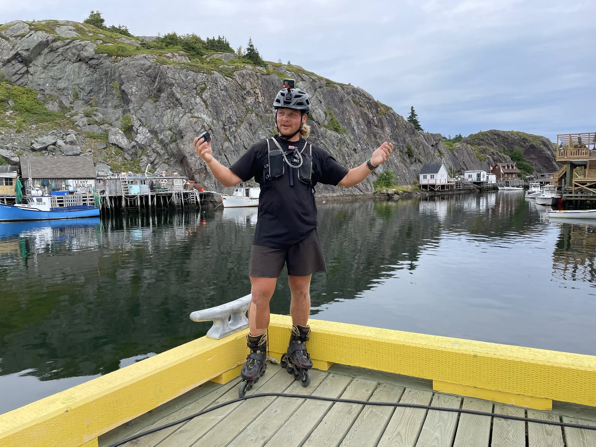 A man wearing rollerblades, a black helmet with a GoPro camera, and a black t-shirt stands on a yellow dock by the water, holding a small device in one hand. The background features a scenic harbor with boats, small houses on stilts, rocky hills, and a cloudy sky.
