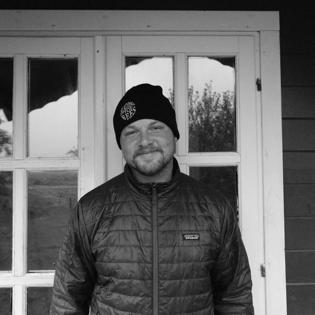 Black and white photo of a man smiling, wearing a knit beanie and a quilted jacket, standing in front of a window with trees visible outside.