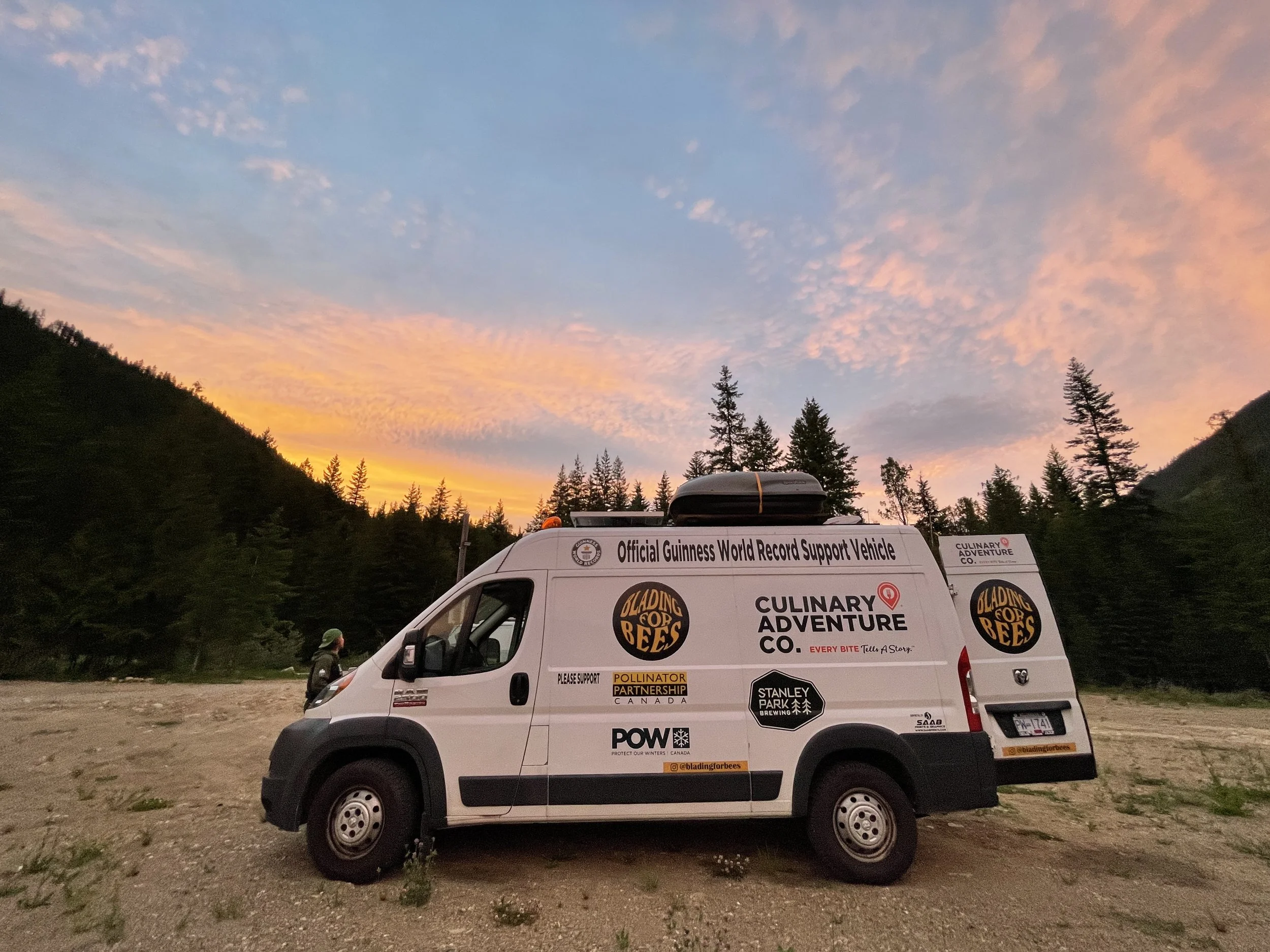 A van parked in an open area with a mountain and pine trees in the background during sunset. The van has various stickers and logos, including 'Official Guinness World Record Support Vehicle' and 'Culinary Adventure Co.'