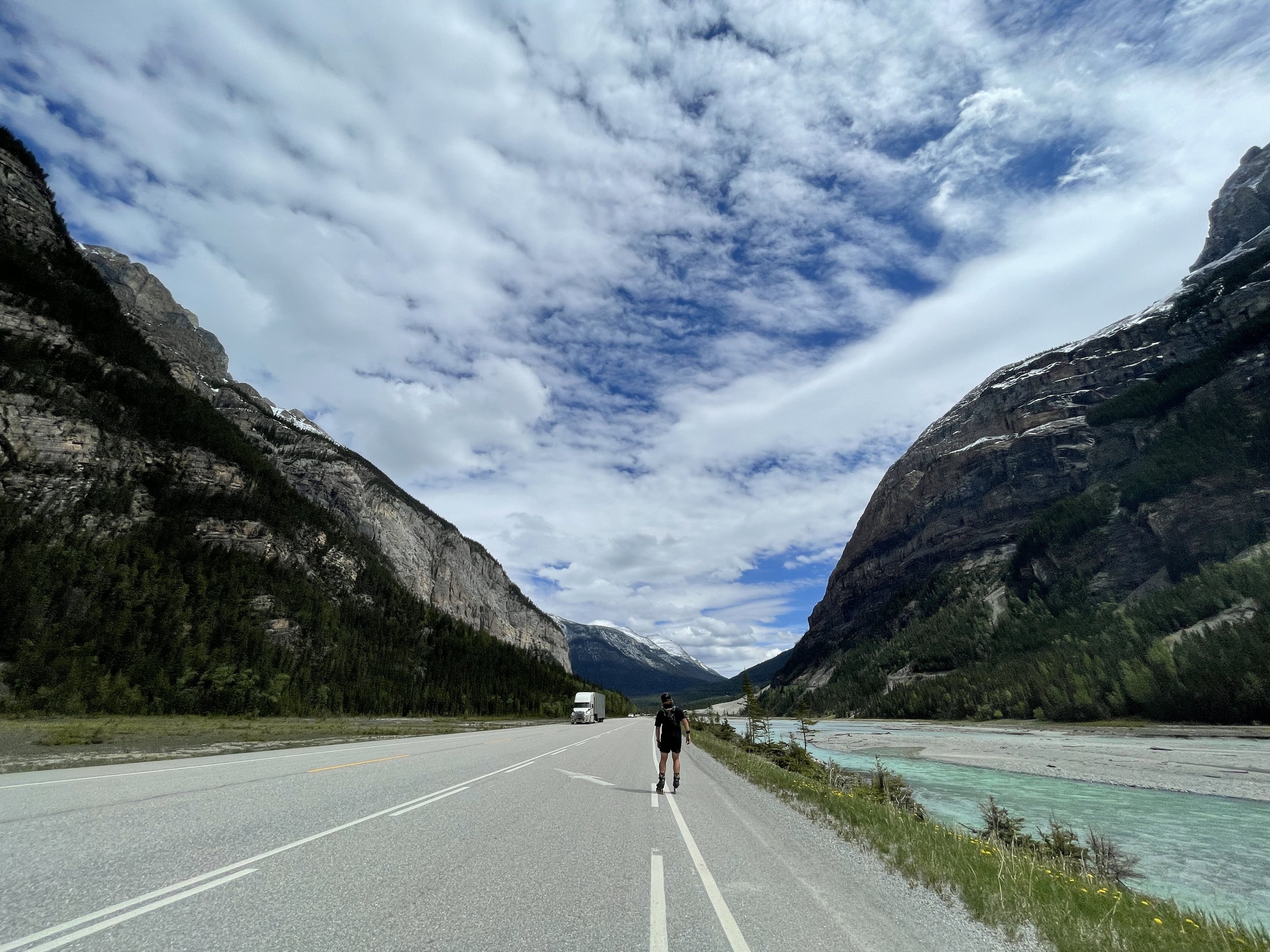 Person rollerblading on the side of a scenic mountain road with towering mountains on either side, a river to the right, and a sky filled with clouds overhead.